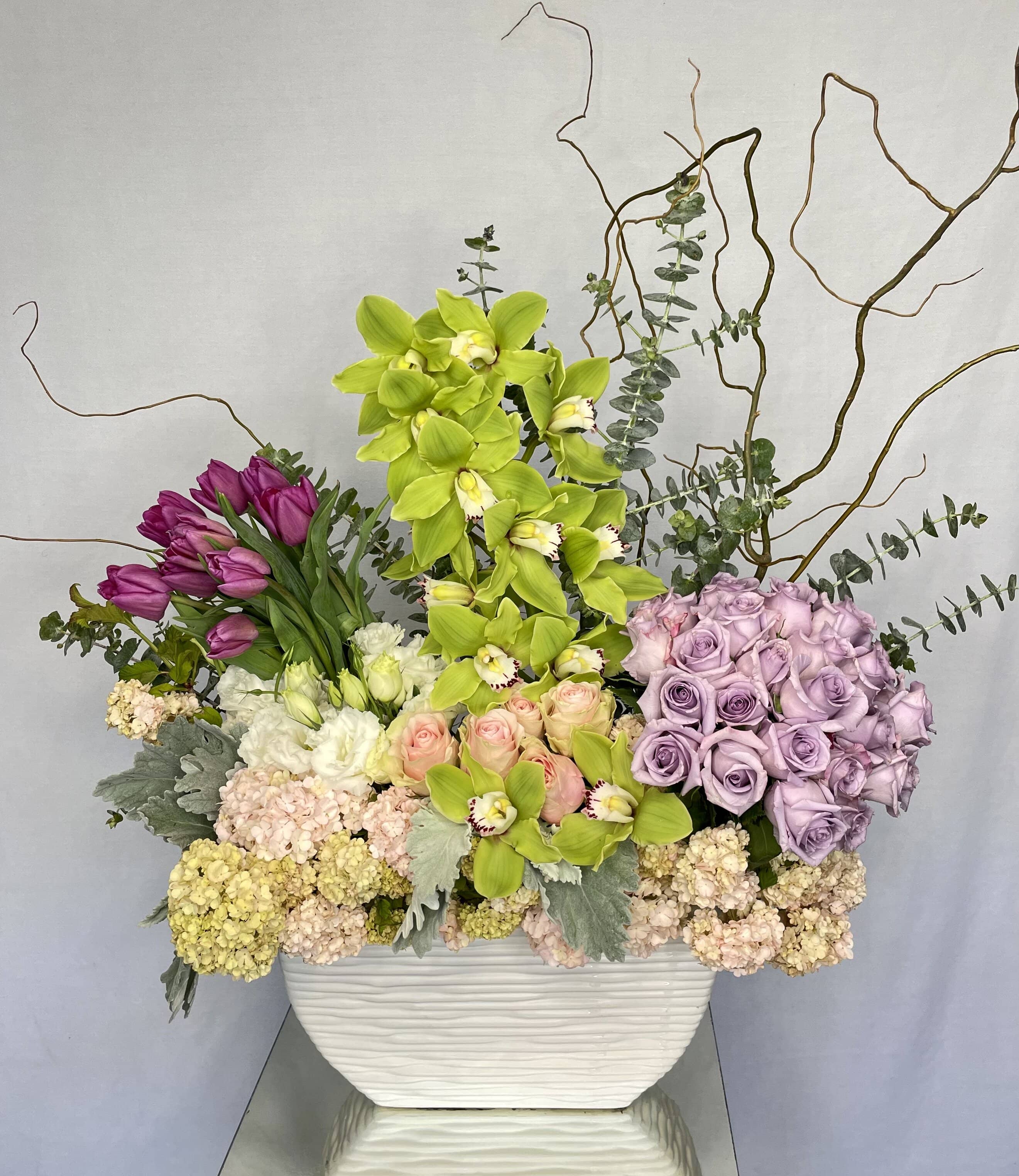 Modern Show - A large beautiful design featuring Green Cymbidium Orchids, Purple Tulips, Light Pink Roses, White Spray Roses, Lavender Roses, Pink Hydrangeas, Dusty Miller, Curly Willow, in a large white vase