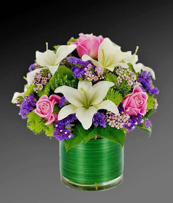 Loving Lily - There simply are not enough superlatives to adequately describe this floral masterpiece. White Asiatic lilies, pink roses, purple statice, green poms, green trachelium and pink waxflower create floral magnificence in a clear vase with Ti leaf ribbon. Measures 12&quot;H by 12&quot;L.