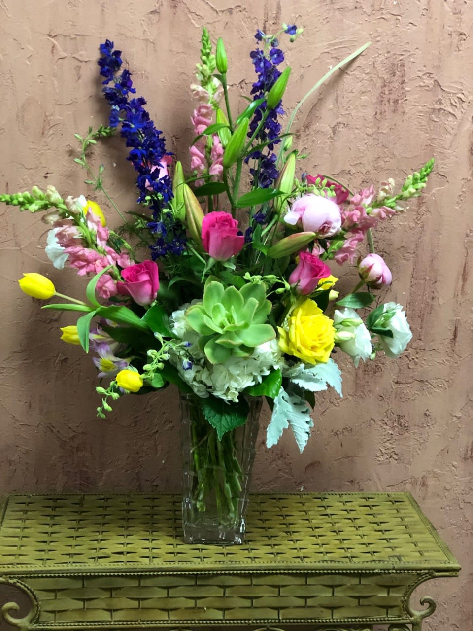 June Bloom - Type of Flowers: Pink Snapdragon, Purple Larkspur, Pink Peonies (seasonal), Pink Stargazers, Yellow Roses, Pink Roses, White Hydrangeas, White Lisianthus, Yellow Tulips, Green Succulent, and Dusty Miller in a short vase. Availability: Seasonal  Peonies available November-March, May-June.  Substitute Available: Yes Design View: Symmetric Front Facing View Photo shown: Regular