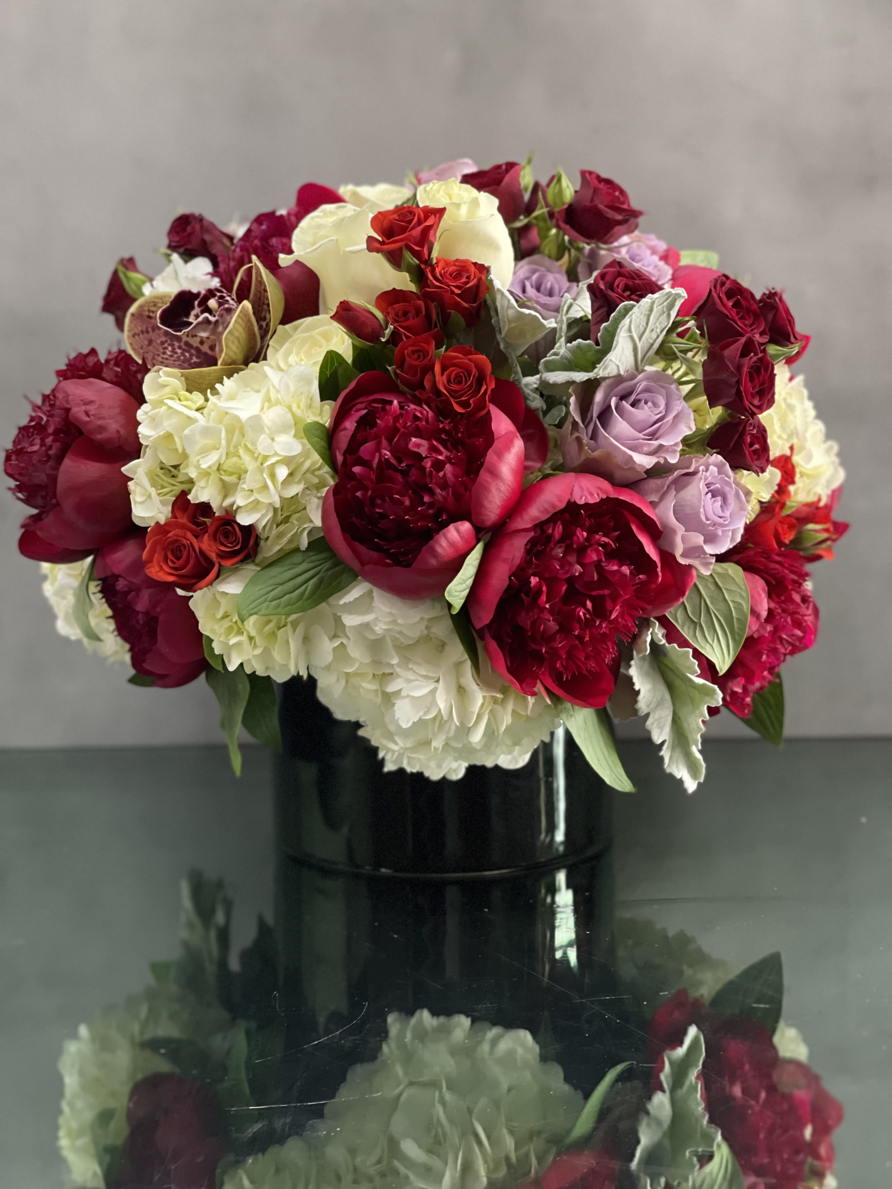 Color My World - This arrangement includes peonies, lavender and red roses, cym orchids , nested on white hydrangeas in a black cylinder vase. A stunning decoration for any home or office. 