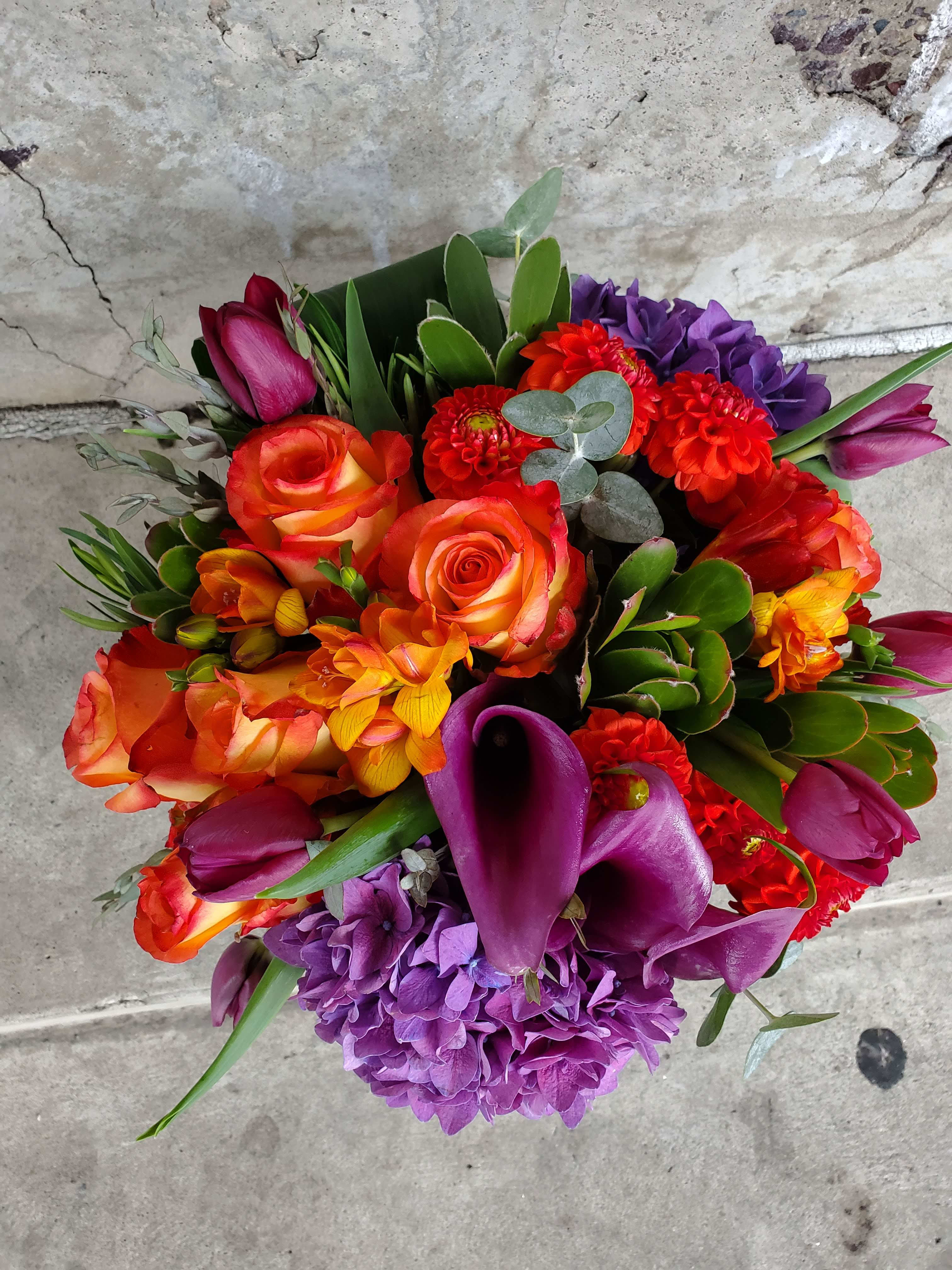 Awaking - An organic explosion of colors mix with hydrangea, purple callas, tulips and vibrant orange roses, with a light accent of organic greens in a clear vase.