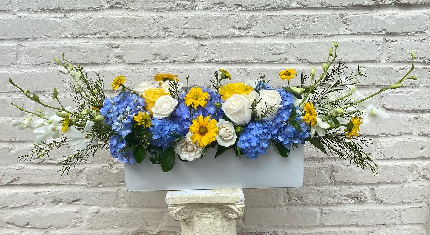 Blue Centerpiece - Arrangement of blue hydrangea  and white roses, daisies, accented with fresh greenery. Artistically designed by our florists in a white container.