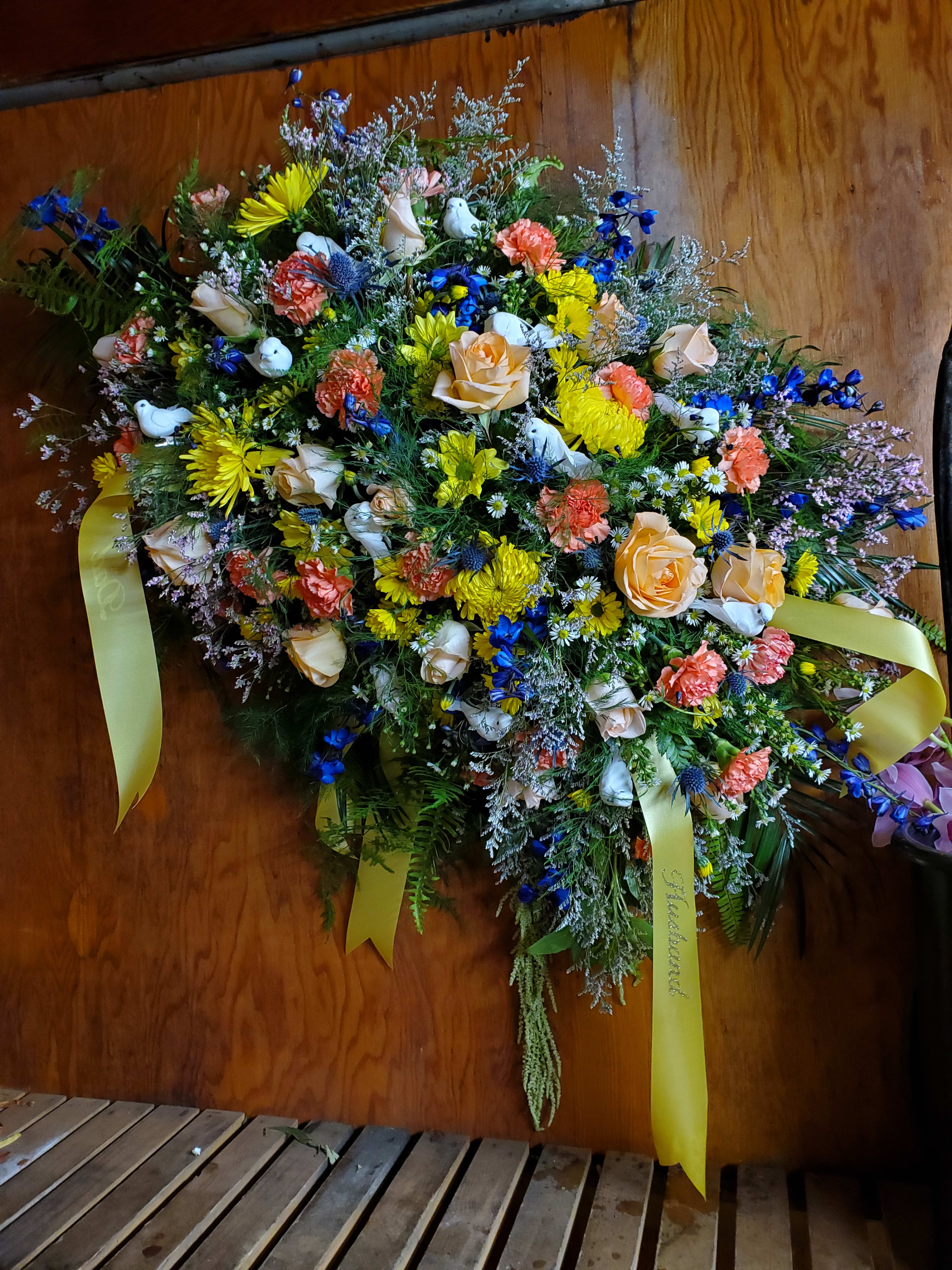 Dovely Casket Spray - Wide variety of flowers throughout spray, including Roses, Thistle, Blue Delph, Carns, Vikings, and more, with fillers, greens and accented with white doves  225.00-300.00