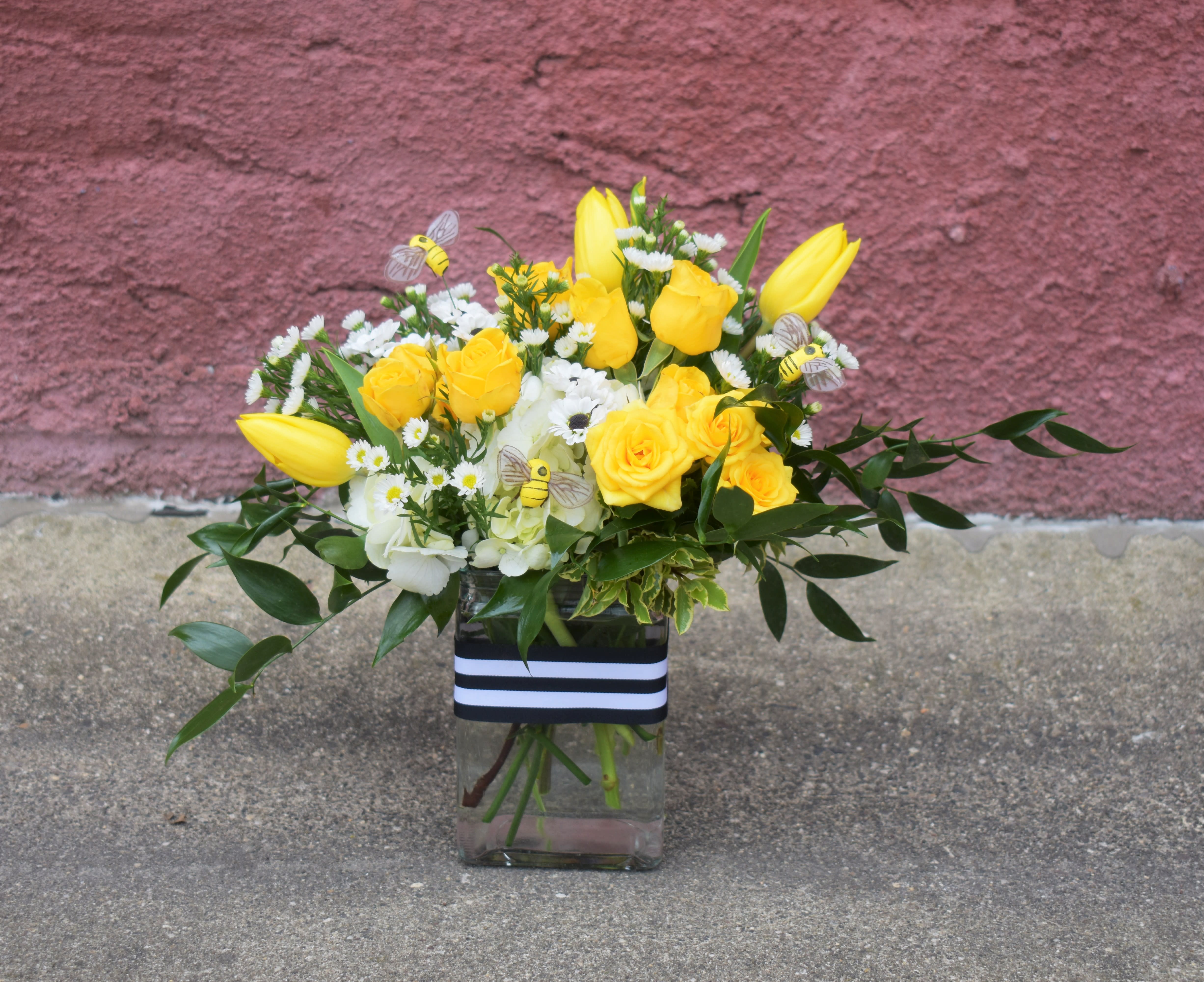 Honey Bee - This adorable bouquet is designed in a glass vase with yellow tulips, yellow spray roses, white hydrangea, white asters, lush greenery, and accented with cute little bees and a black/white striped ribbon band. A fun and cheery gift for any occasion! . 