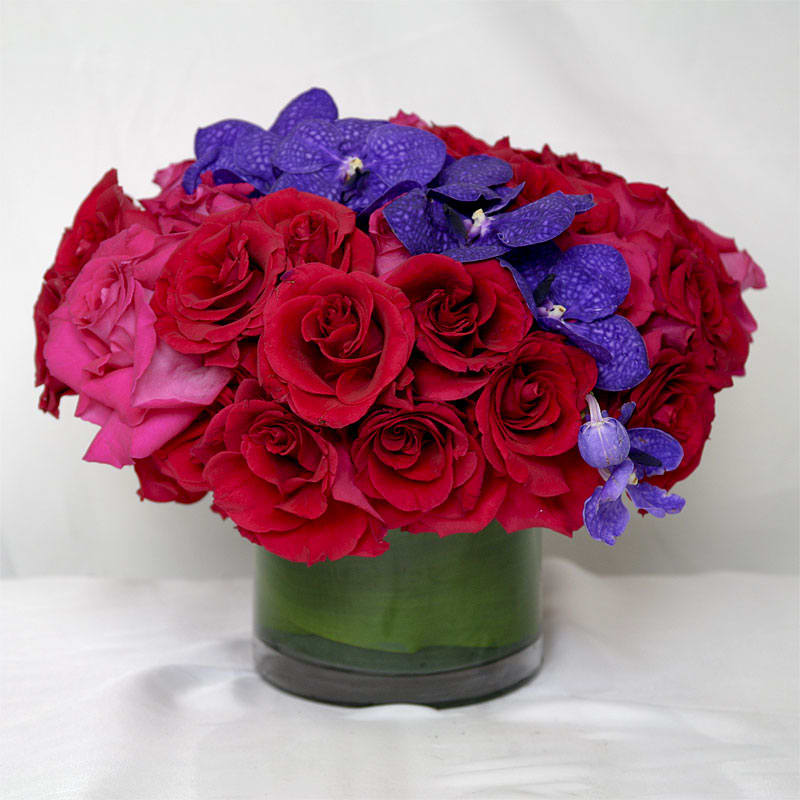 Scarlett O'Hara Scarlett O'Hara - Red and magenta roses with a full stem of purple vanda orchids in a circular glass vase lined with aspidistra leaves.