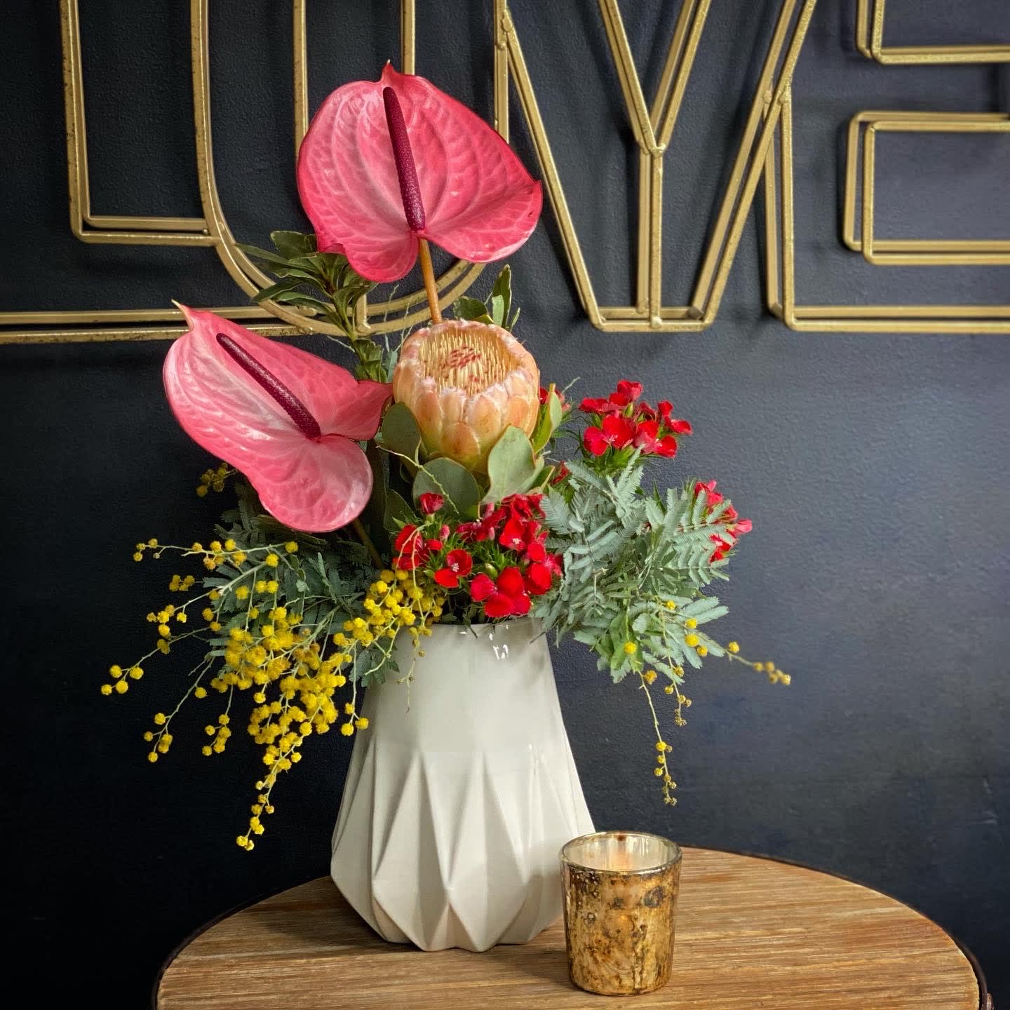 Keep it HOT! - This tropical arrangement is like a heat wave in a vase! It will instantly provide a vacation vibe wherever it goes. It includes interesting tropical elements like protea and anthurium in a modern, geometric vase and stands about 12-16&quot; tall. COLORS WILL VARY.