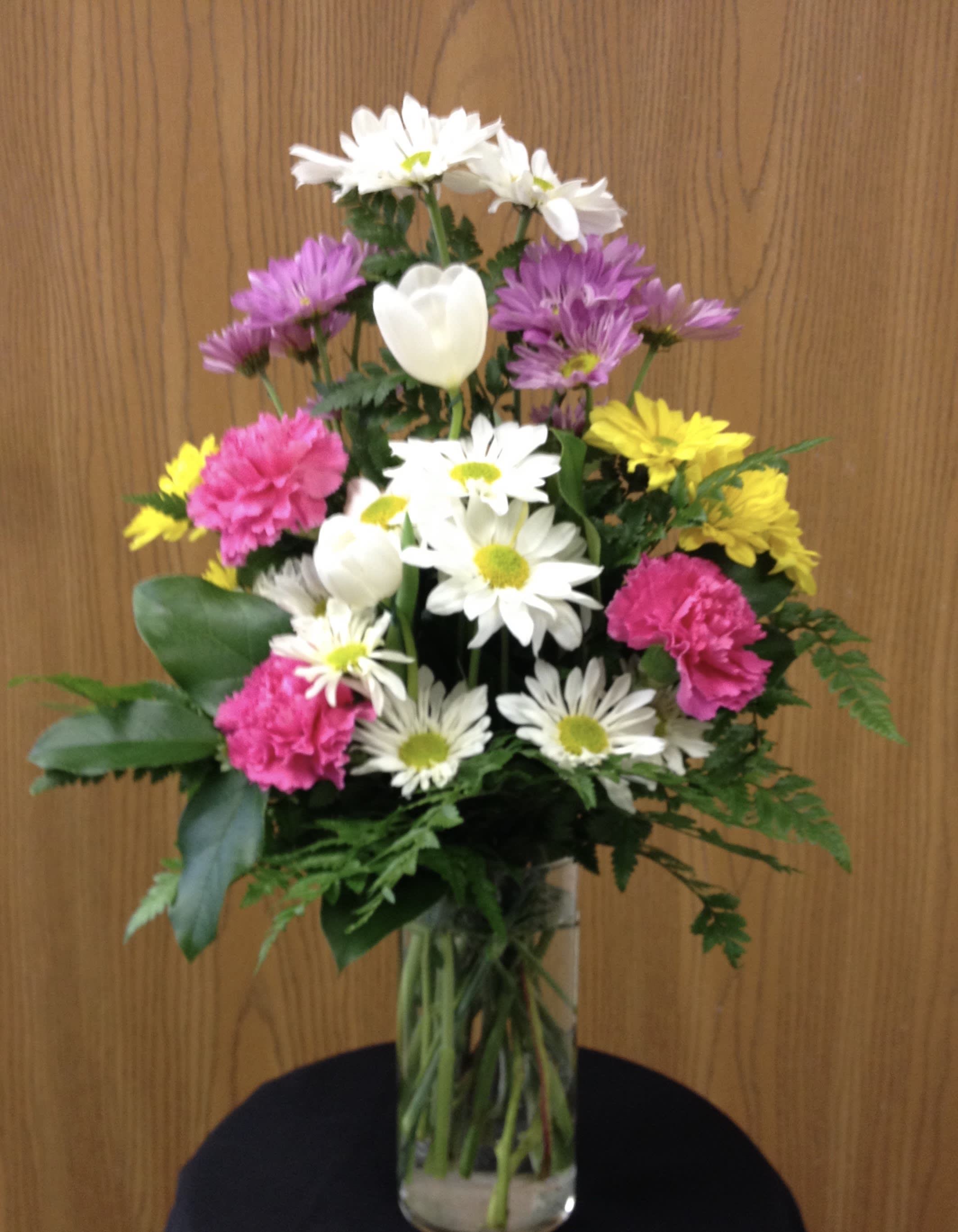Cheerful Blossoms - A bright assortment of flowers SUCH AS carnations, daisies, and tulips with assorted greenery in a clear vase.
