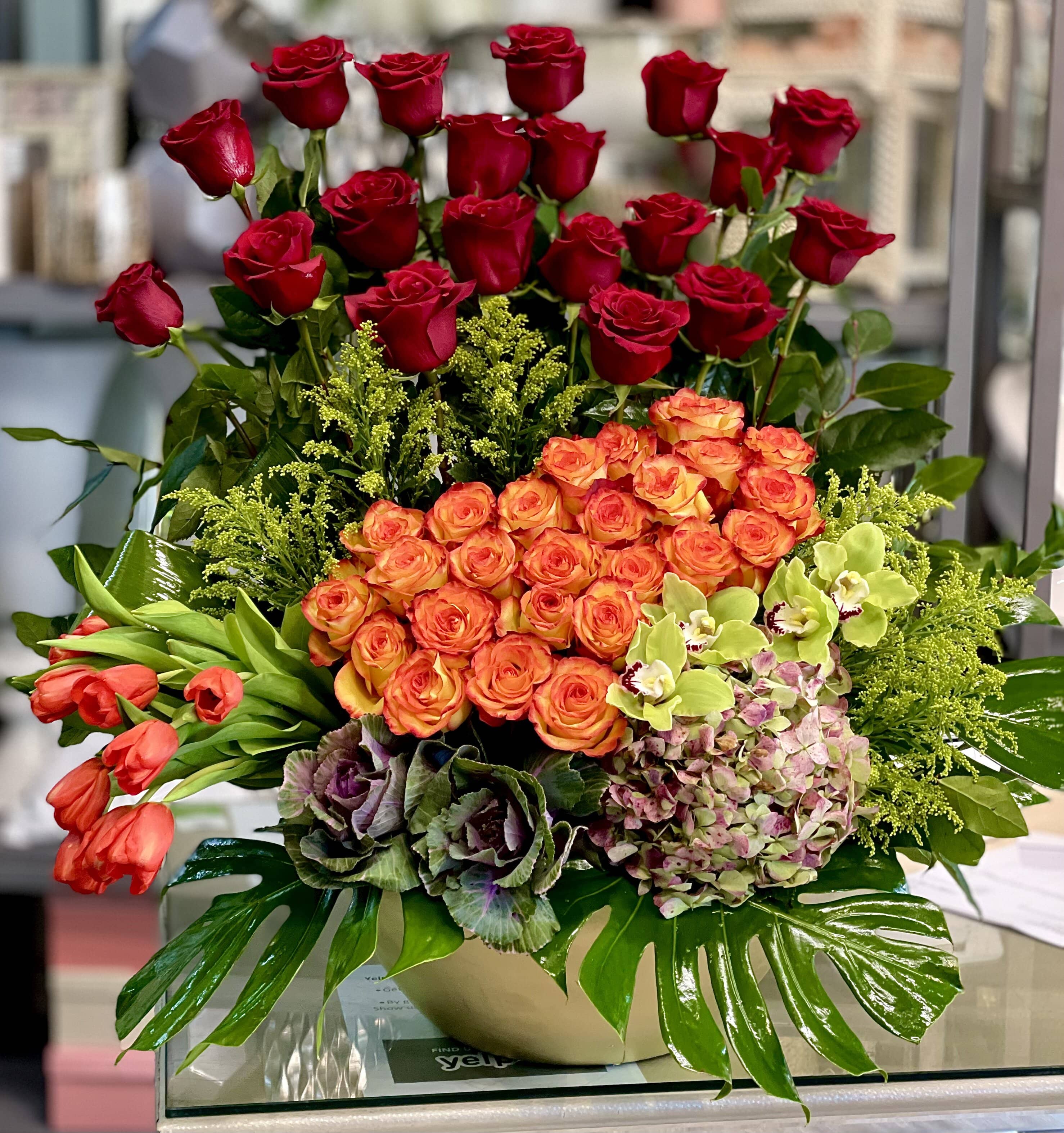 The Wildest Dreams  - Red Roses, Orange Roses, Green Cymbidium Orchids, Orange Tulips, Hydrangeas, and Monstera Leaves in a large round vase.