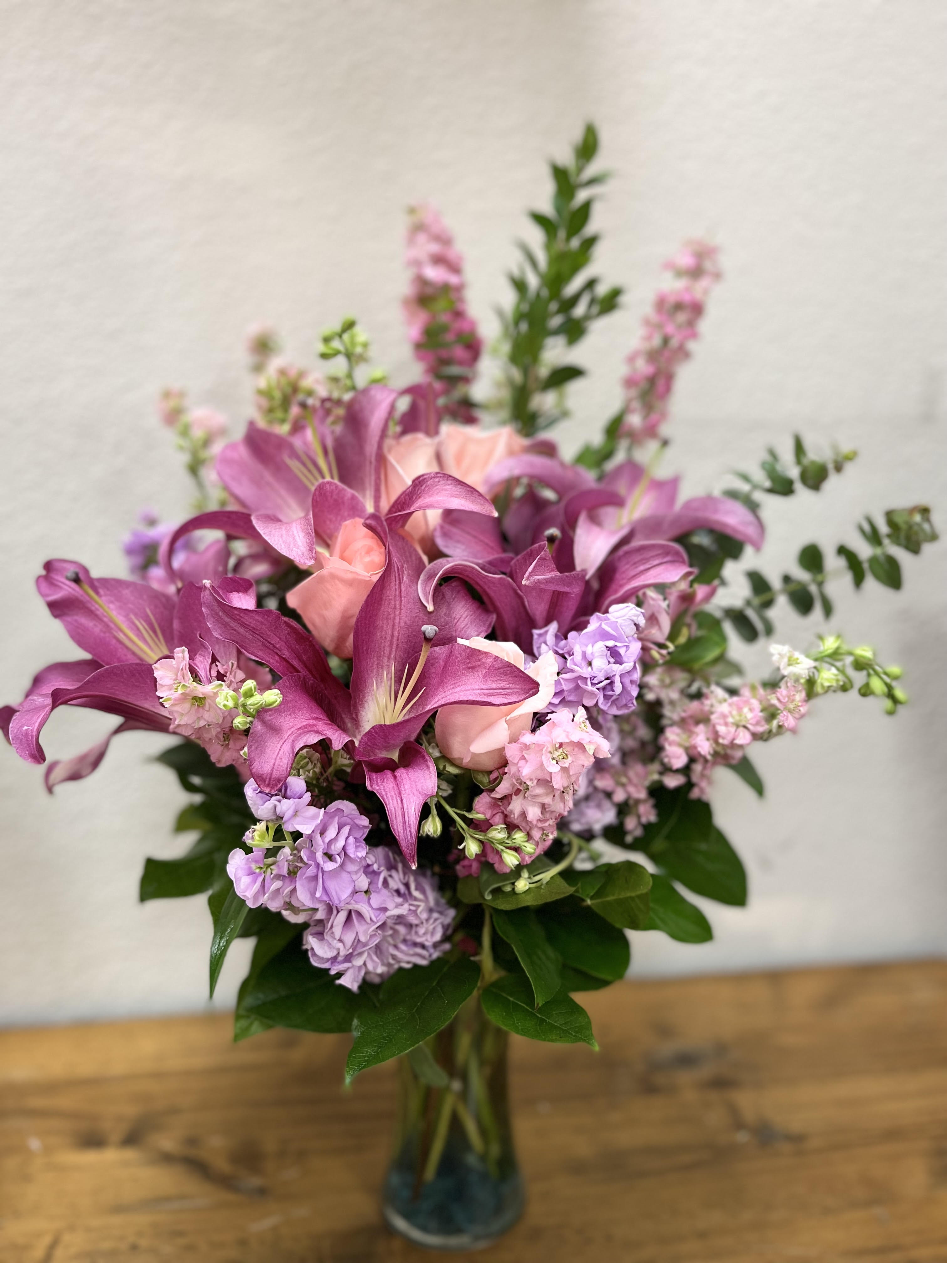 Soft Blooms Petals - A touching tribute to a beloved woman, this beautiful, soft pink and lavender arrangement is presented in a large antiqued pot for an elegant touch. This soft pink arrangement features lavender roses, pink spray roses, pink asiatic lilies, lavender carnations, pink larkspur, queen anne's lace, huckleberry, dusty miller, and leatherleaf fern. Delivered in a Classical Garlands pot.   