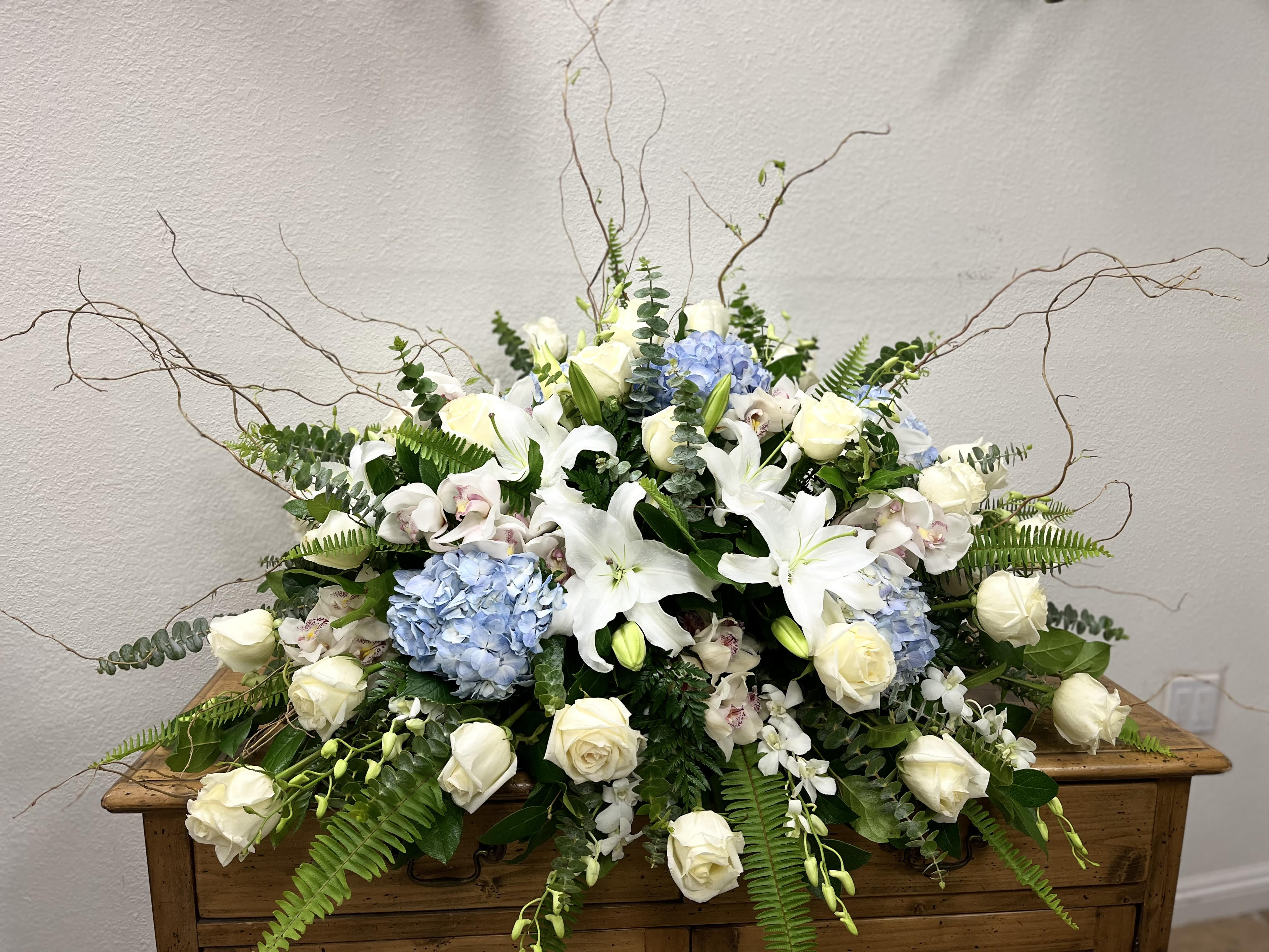Ocean Mist Casket Spray - As serene as gently falling snow, this pure white spray of hydrangea, orchids, roses and lilies is a heartfelt symbol of peace and beauty--a symbol that will remain a guiding light to your loved ones for years to come. This stunning spray features blue hydrangea, white cymbidium orchids, white dendrobium orchids, white roses, white spray roses, white oriental lilies, curly willow, green ivy, parvifolia eucalyptus, seeded eucalyptus, silver dollar eucalyptus, sword fern, and lemon leaf.