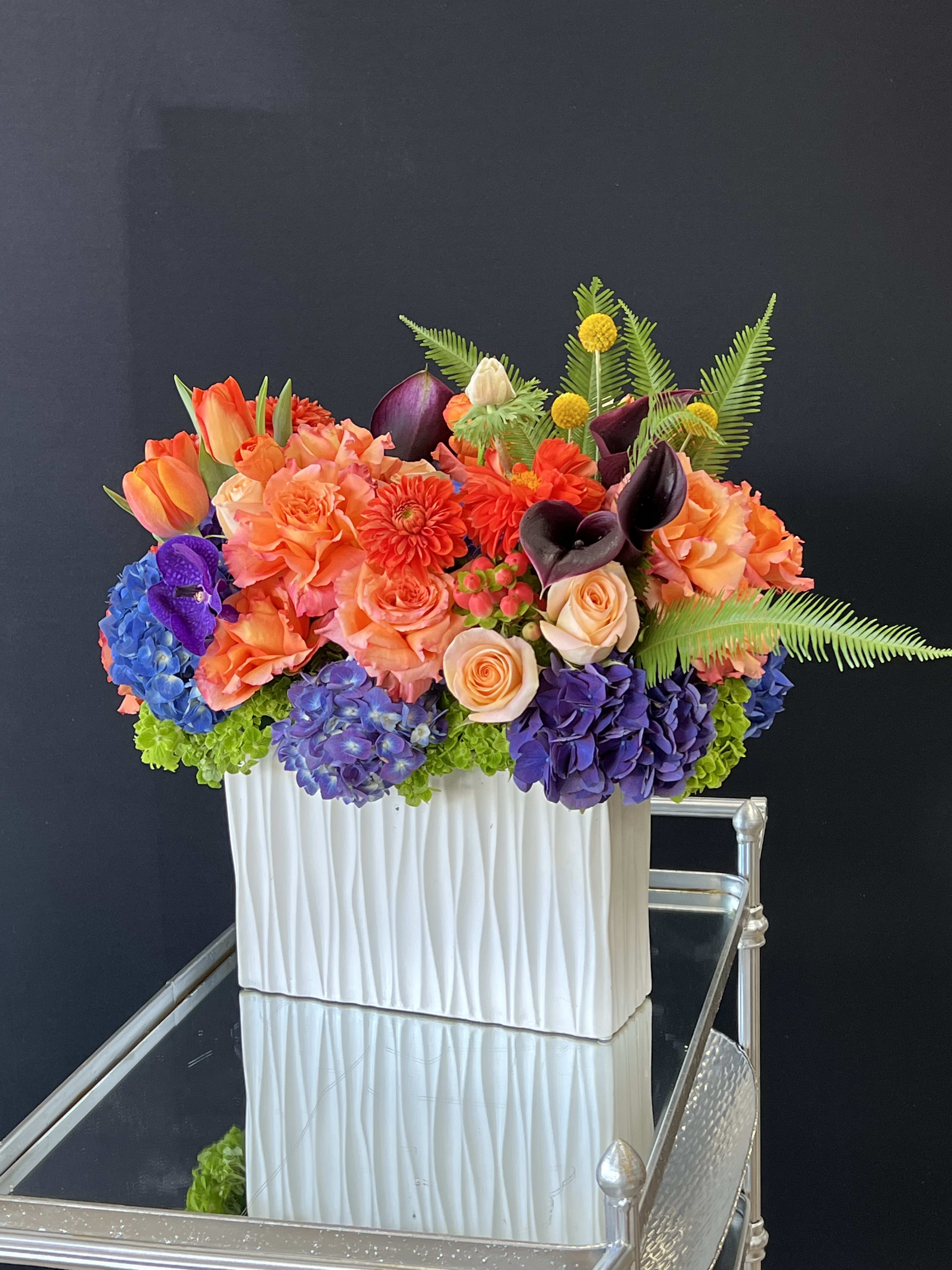 Aztec Beauty - Orange Ranunculus, Purple orchids, magenta dhalias, Lilacs and Yellow tulips in a beautiful oblong vase