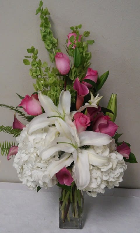 Roses Lilies and Hydrangea Roses Lilies and Hydrangea - White Hydranga and Lilies with Pink Roses, Cala Lilies, Bells of Ireland and Greenery in a tall Square Cylinder Vase.