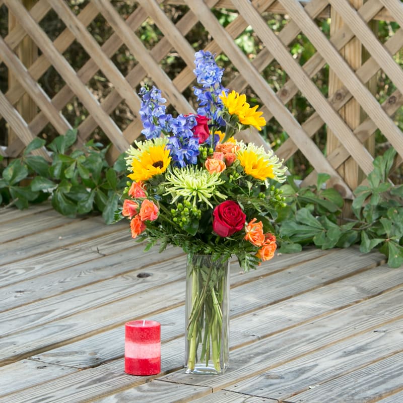 Sweetly Popping - Sweet arrangement with sunflowers, orange spray roses, blue delphinium, green spider mums, and red roses. 