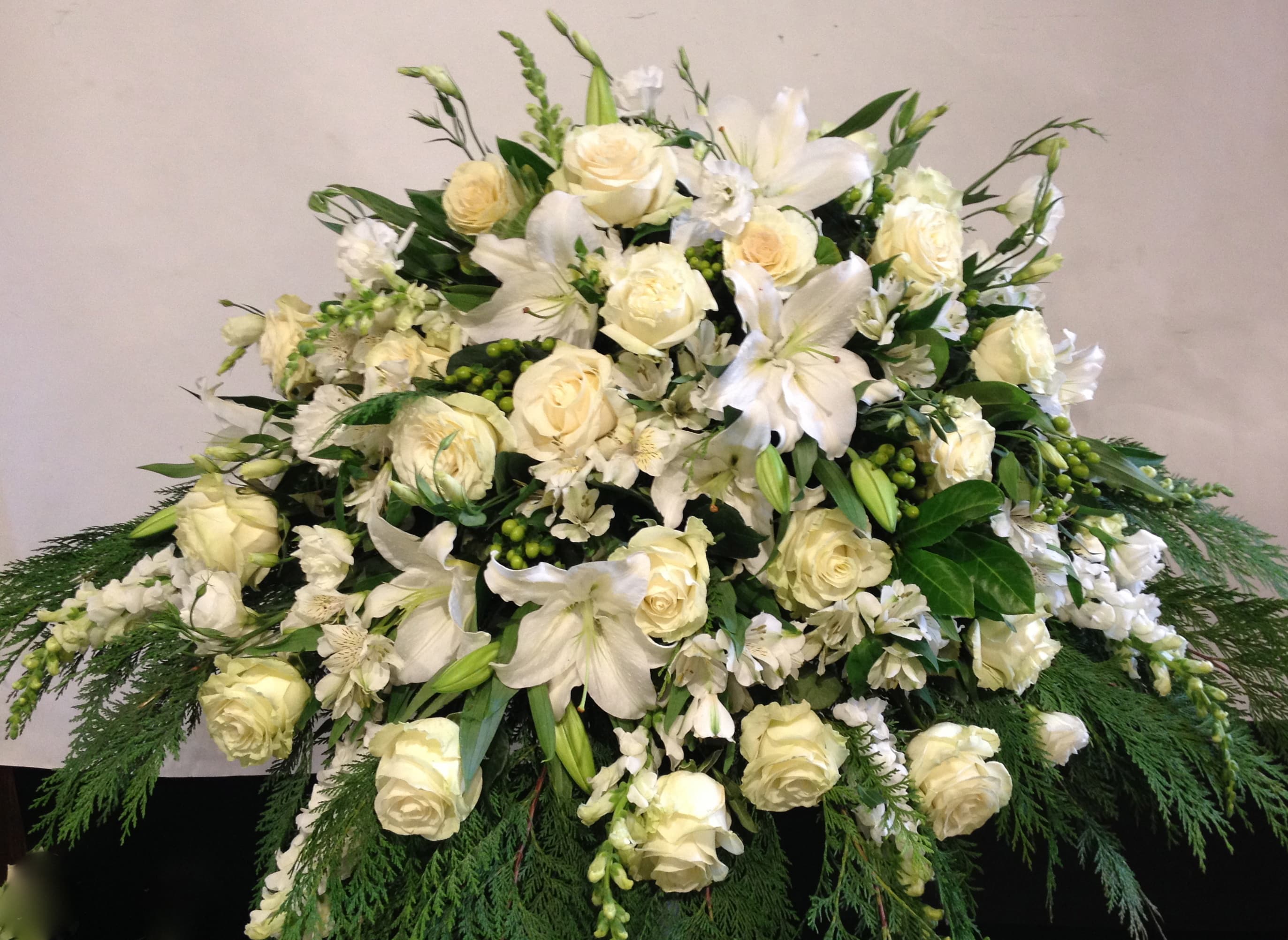 Pure White Casket Blanket  - This casket blanket inspires thoughts of comfort and peace to those wishing to pay their respects for the loss of the deceased. White roses, lilies, snapdragon and other seasonal flowers are accented with an assortment of the finest lush greens to create the perfect arrangement to display on the top of their casket during their final farewell service.