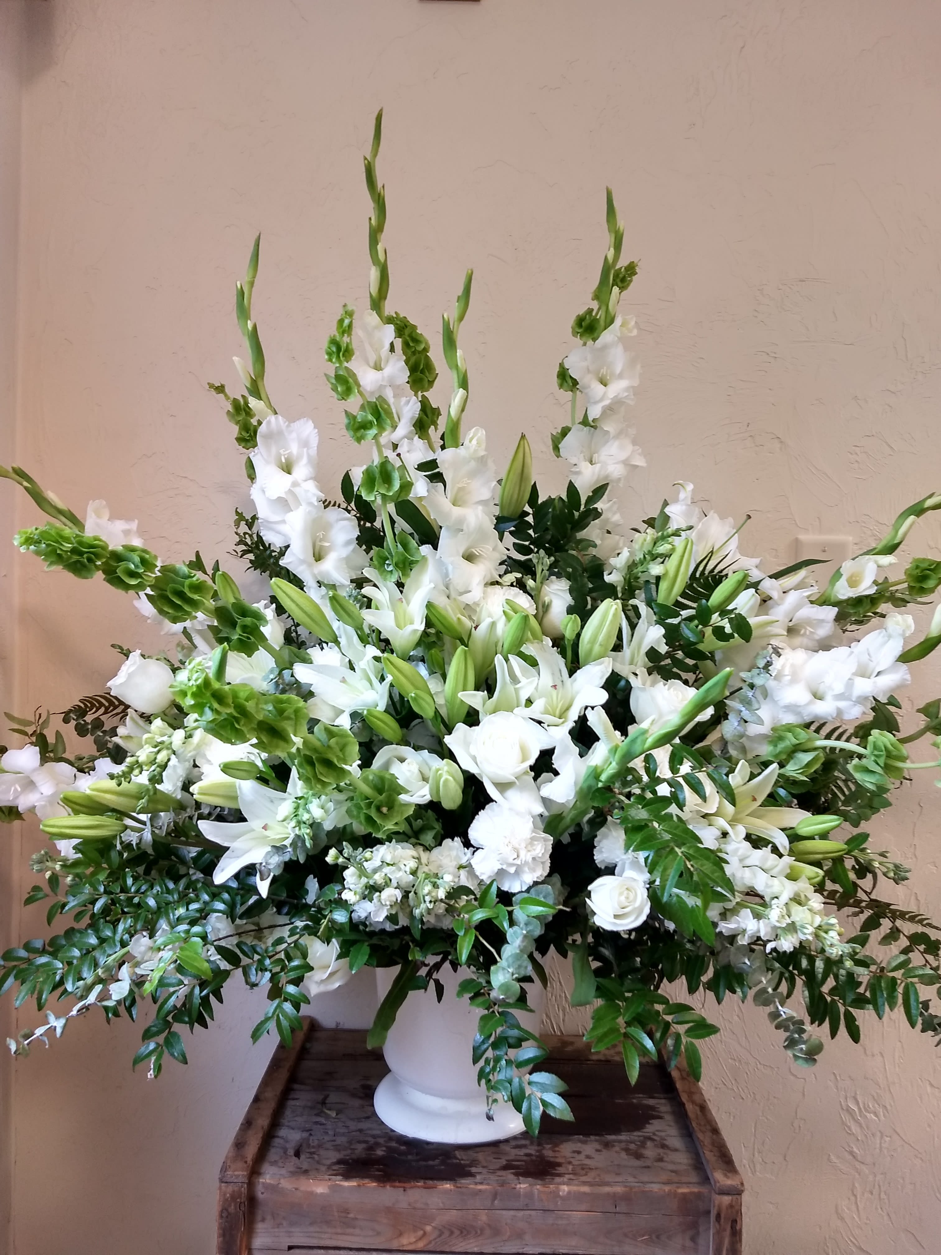 Rising Star by Cricket's Flowers cfS24438p - This sympthy arrangement is a brilliant expression of peace and soft serenity. White roses, carnations, gladiolus, stock and Oriental lilies are accented with stems of Bells of Ireland and an assortment of lush greens, in a white urn.  **** Please note: 48 hour notice MAYBE REQUIRED We custom-design this arrangement using best-of-day seasonal flowers. This image is a general representation of its size and style and may not feature the exact flowers shown. Due to disruptions in the global supply chain and staff shortages, we may have to make last minute substitutions to your selected design. If we need to make significant changes we will call you first to get your approval*** Thank you for understanding.