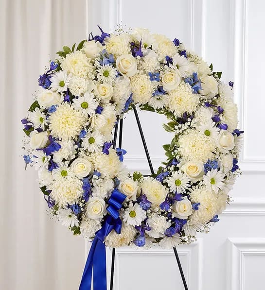 Serene Standing Wreath - Blue and White - Helping someone find peace after the loss of a loved one can be difficult. If you want to offer your condolences through flowers, our timeless standing wreath in blue and white is a beautiful choice. Crafted with a mix of soothing blooms for a lush, full presentation, this touching tribute helps you express all the love and support you have in your heart when they need it most.  One-sided standing wreath arrangement with white roses, football mums, daisy poms and carnations, shades of dark and light blue delphinium; accented with baby’s breath, assorted greenery and blue ribbon. Appropriate for the funeral home. Our florists hand-design each arrangement, so colors and varieties may vary based on local availability.