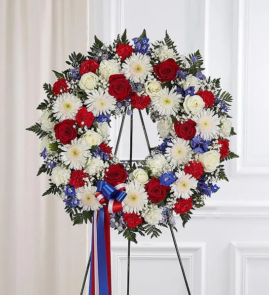 Serene Standing Wreath- Red, White &amp; Blue - For the Service - They served their country with honor and pride, so it’s only fitting to honor them with a beautiful symbol of eternal life. Our patriotic standing wreath arrangement is meticulously crafted by our expert florists to honor a brave veteran who has passed away. Filled with lush blooms in red, white and blue, it’s a fitting final tribute for the funeral services.  Standing wreath arrangement with red roses, and carnations; white roses, cremones and carnations; blue delphinium; accented with baby’s breath and soft lush greenery. Appropriate for the funeral home. Our florists hand-design each arrangement, so colors and assortment may vary based on local availability.