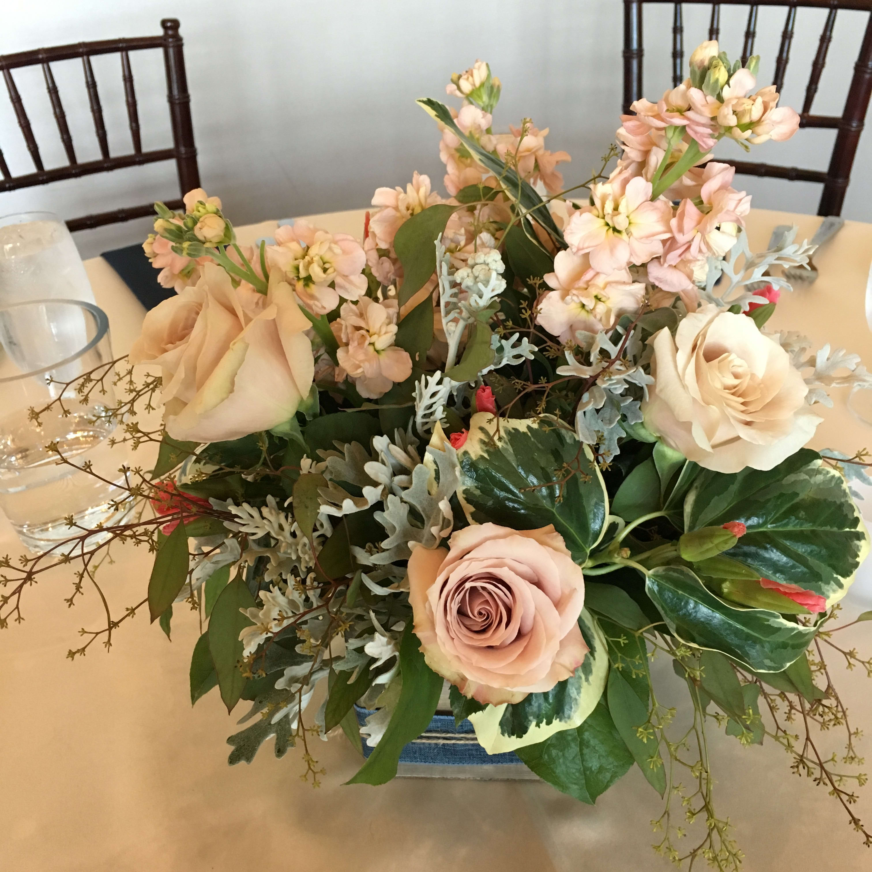 Rustic Wooden Box Rose Centerpiece Rustic Wooden Box Rose Centerpiece - Elegant creation with a few roses, seasonal flowers and premium greenery. This bouquet is designed in a simple wooden box with burlap ribbon tied around it. Vintage style! Selection & color scheme will vary. Choose your own colors when you order at least one week ahead. Suitable for a round or square dining table of up to five guests, this is a low centerpieces with flowers all the way around.