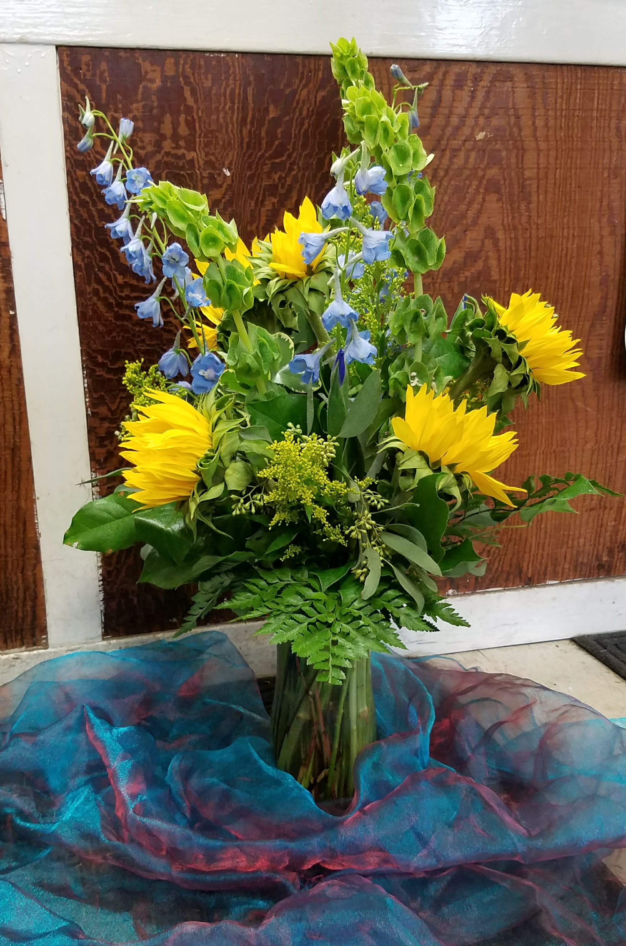 Sunshine Sky - This cheery arrangement is a great way to brighten up any space. Sky blue delphinium and Sunflowers are the stand-out blooms here. Green Bells of Ireland add contrast, and blue iris make the finishing touch.