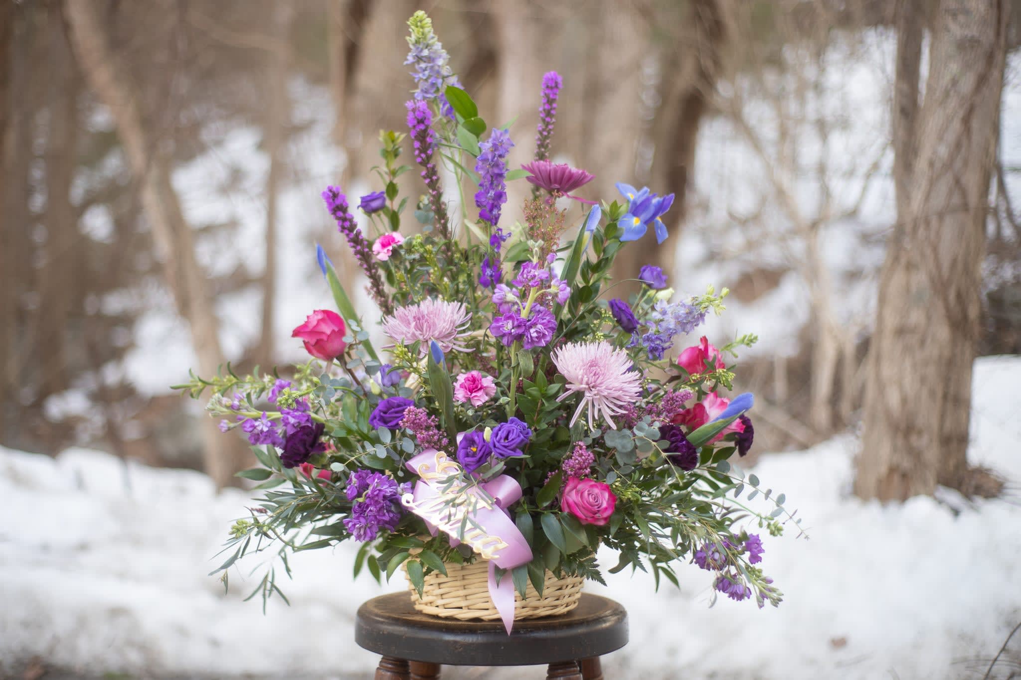 Lavender Garden Basket - Wildflower inspired floral tribute basket in lovely lavender, indigo and purple tones traditionally designed in a one sided fashion to display beautifully in the funeral parlor or at a graveside service. Tall and airy larkspur blooms in a violet hue embrace the garden spirit alongside towering liatris blooms. Focal flowers of roses and cremons create a graceful display of love and admiration. Metallic gold script is available for all floral memorial tribute pieces. Basket styles and colors may vary from photo. Standard price is displayed in photo. Can be created in a multitude of color palettes if desired. 
