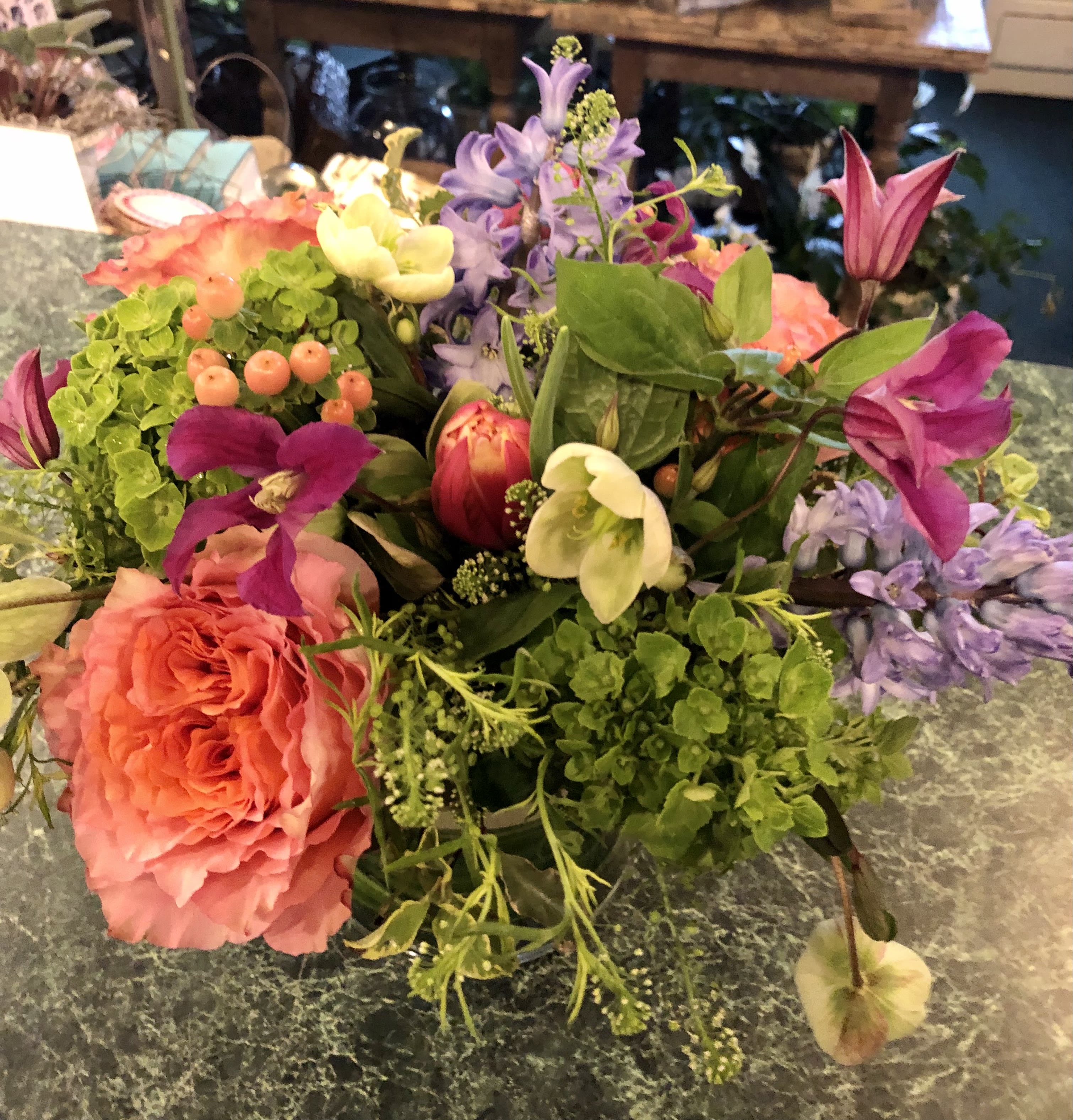 Free Spirit Spring Cylinder - Mix of Free Spirit roses, green hydrangea, hellebores, blue hyacinths, Columbus tulips, peach hypericum and Kiev clematis in 5x5 cylinder vase leaf lined with lepidium and  variegated mini pittisporum.