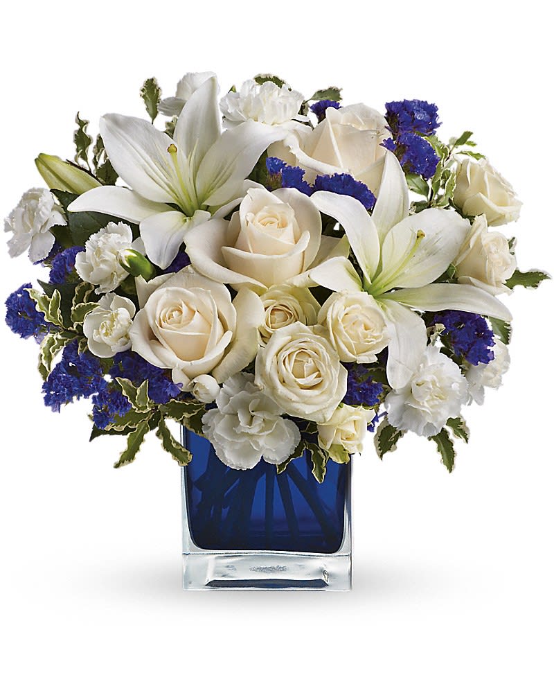 Diamonds in the Sky - Send someone a bit of heaven with this beautiful bouquet. Luxurious crème roses and pure white lilies paint a peaceful picture inside a sapphire blue cube. Crème roses, white asiatic lilies and white miniature carnations are mixed with bursts of purple statice and green pitta negra. Delivered in a glass Cube.Approximately 12&quot; W x 11 1/2&quot; H