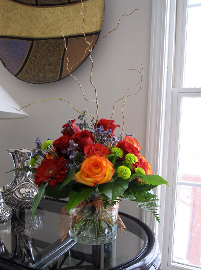 Sunshine - Mainly roses in red and orange colors, some gerbera daisies, accented with mini green mums, limonium and some greenery. Some twigs to give height and a touch from nature. Arranged in a glass cylinder vase, glass beads on the bottom and a metallic ribbon.