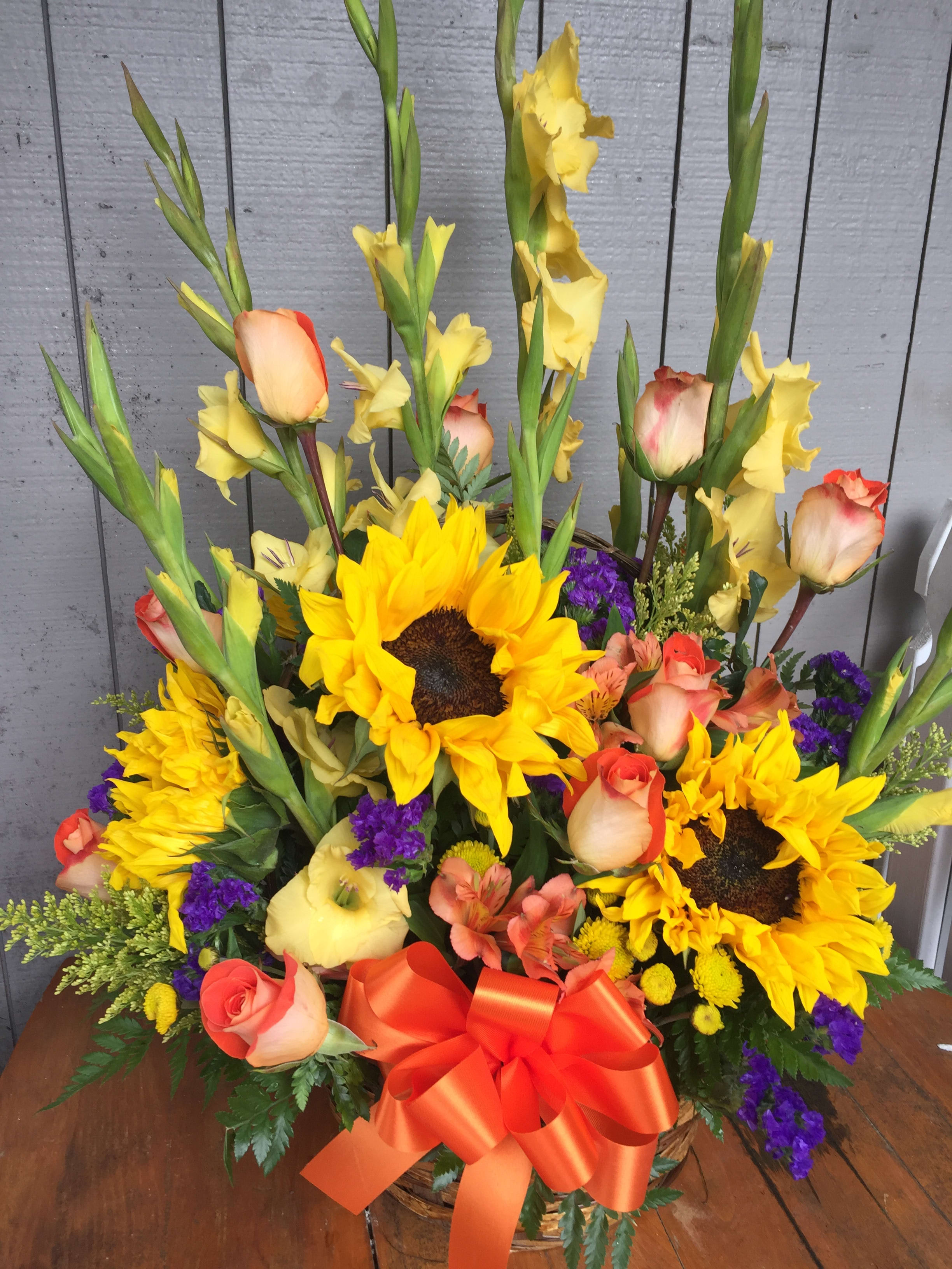 TALL AND SUNNY - This basket arrangement is full of cheer. Yellow gladiolus 10 orange roses, orange alstroemeria, purple status and sunflowers.  Flowers shown have not fully opened.  Approx.  24&quot; tall and  15&quot; across.