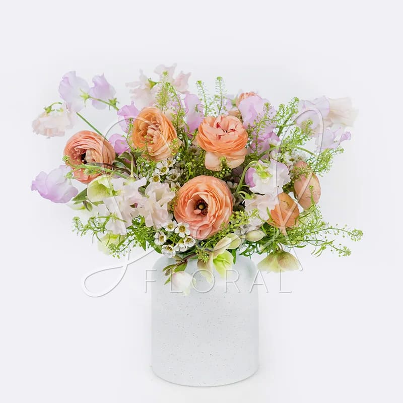 Spring Sweets - A medley of fresh seasonal blooms in shades of peach ranunculus, soft blush and ivories fragrant sweet peas mingling alongside fresh greenery &amp; filler arranged in speckled Flynn Vase 5&quot; x 5.75&quot;. (Arrangement is Full Sided)