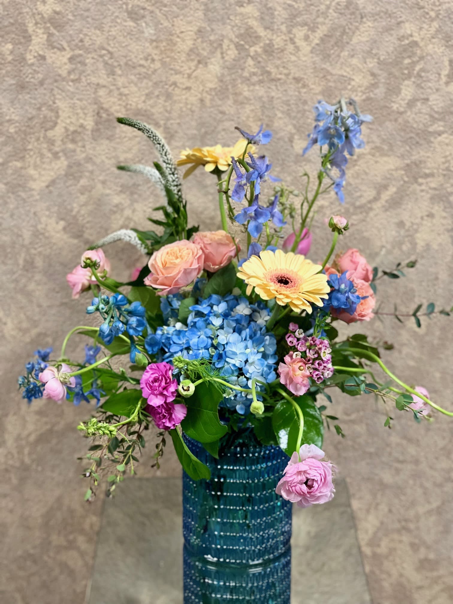 Colorful Soft Hues - This design is great for spring birthdays. Recipe: Soft Hues with Blue Hydrangea, Spray Roses, Larkspur, Ranunculus, and Gerber Daisies in a blue glass vase. Availability: Seasonal Ranuculus Substitute Available: Yes Design View: Symmetric Front Facing View Photo shown: Regular 