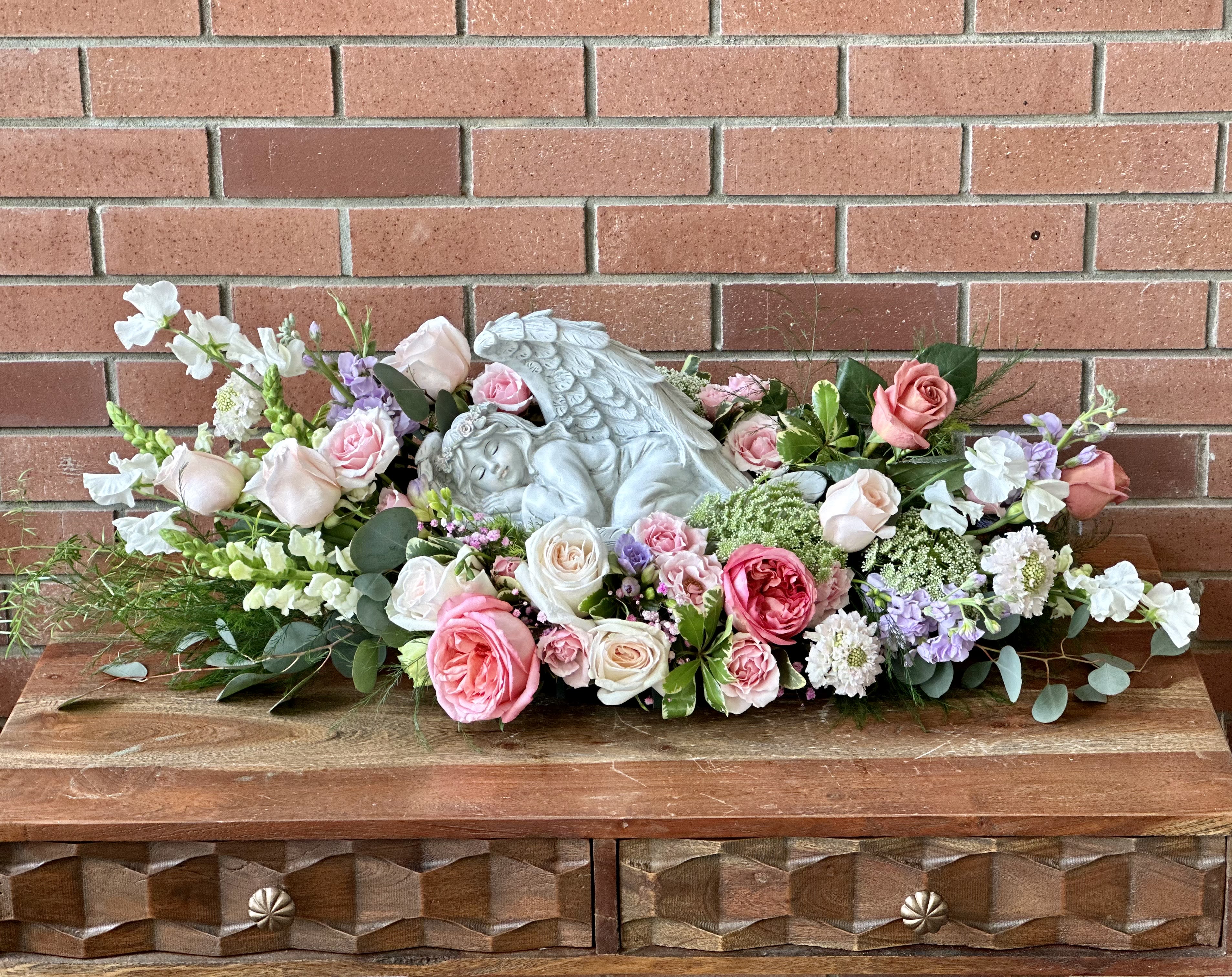 Sleeping Angel Sympathy Arrangement  - Beautiful 13” long sleeping angel surrounded with this weeks’s freshest pink, white and lavender flowers. Roses, garden roses, stock. Spray roses, freesia, queen annes lace, sweet peas and lisianthus arranged among lush greeneries and filler flowers. 