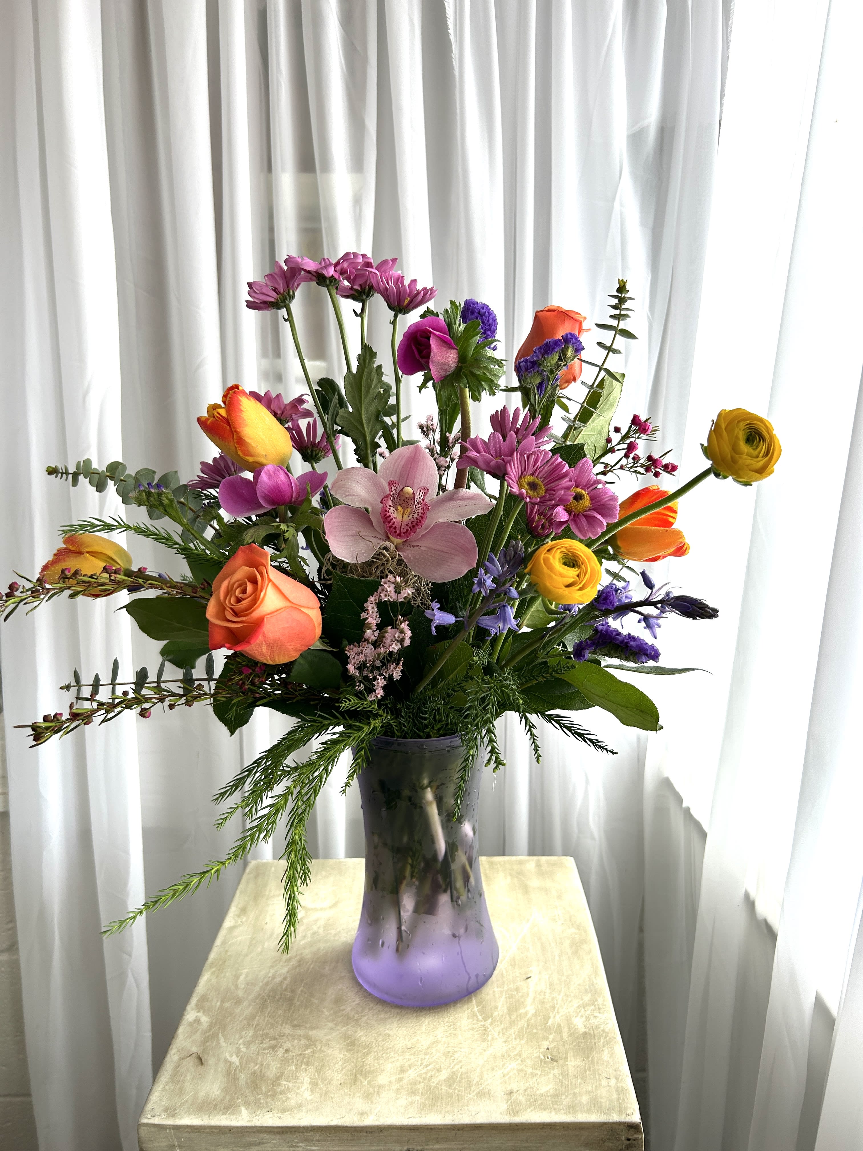 Robin's Rest - A fresh haven of spring.  Anemoneas, ranunculus, tulips, and other wonderful spring flowers gather in a lavender vase.  As always, call shop to find out what's in stock, because our inventory of both flowers and vases constantly changes.