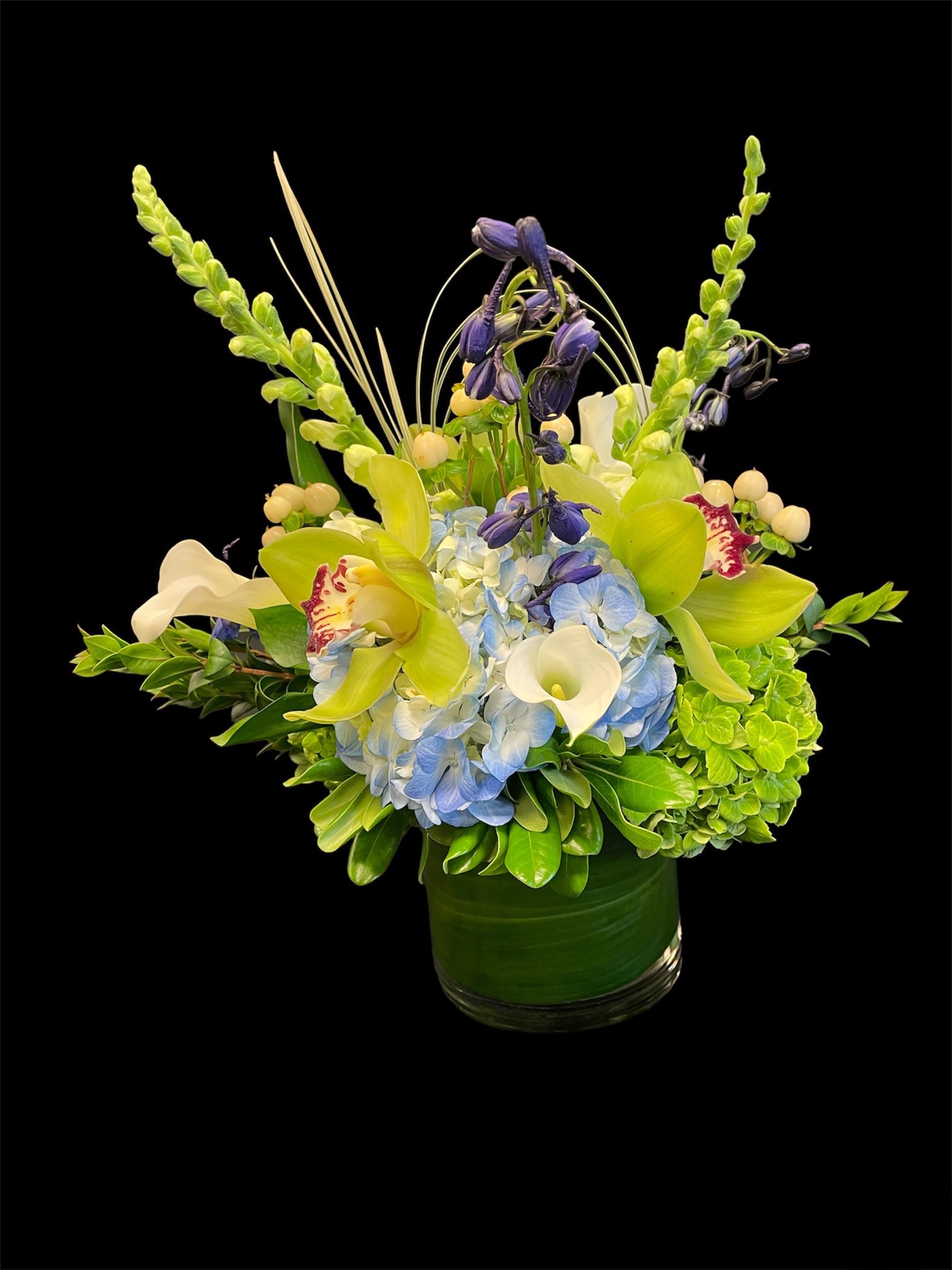 Indoor Jungle - Indoor Jungle includes,  one blue hydrangeas, two green Hydrangeas, three Hypericum Berries, 3 Green Cymbidium Orchid, Two white snopdragons, two blue delphinium and a mixer of greenery. In a 6Hx5W clear cylinder vase with a leaf in the side of it. 