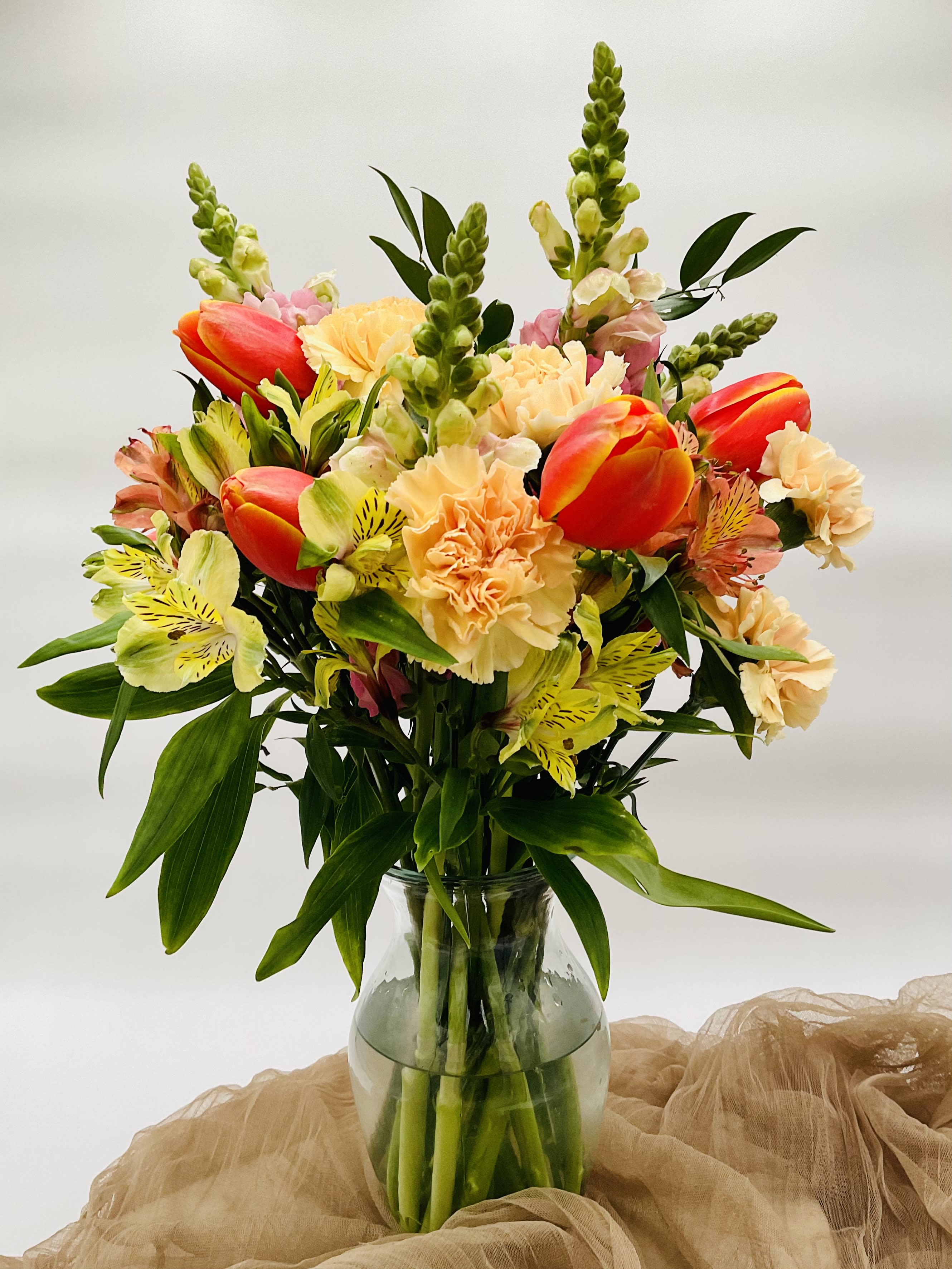Tulips and Cream Bouquet-Mother's Day Tulips and Cream Bouquet-Mother's Day - A peachy and creamy tulip bouquet is perfect for creating dreamy spring memories with your loved ones. Bouquet includes orange-yellow tulips, peach carnations, yellow and pink alstromeria, and pink snapdragons in a clear glass vase.