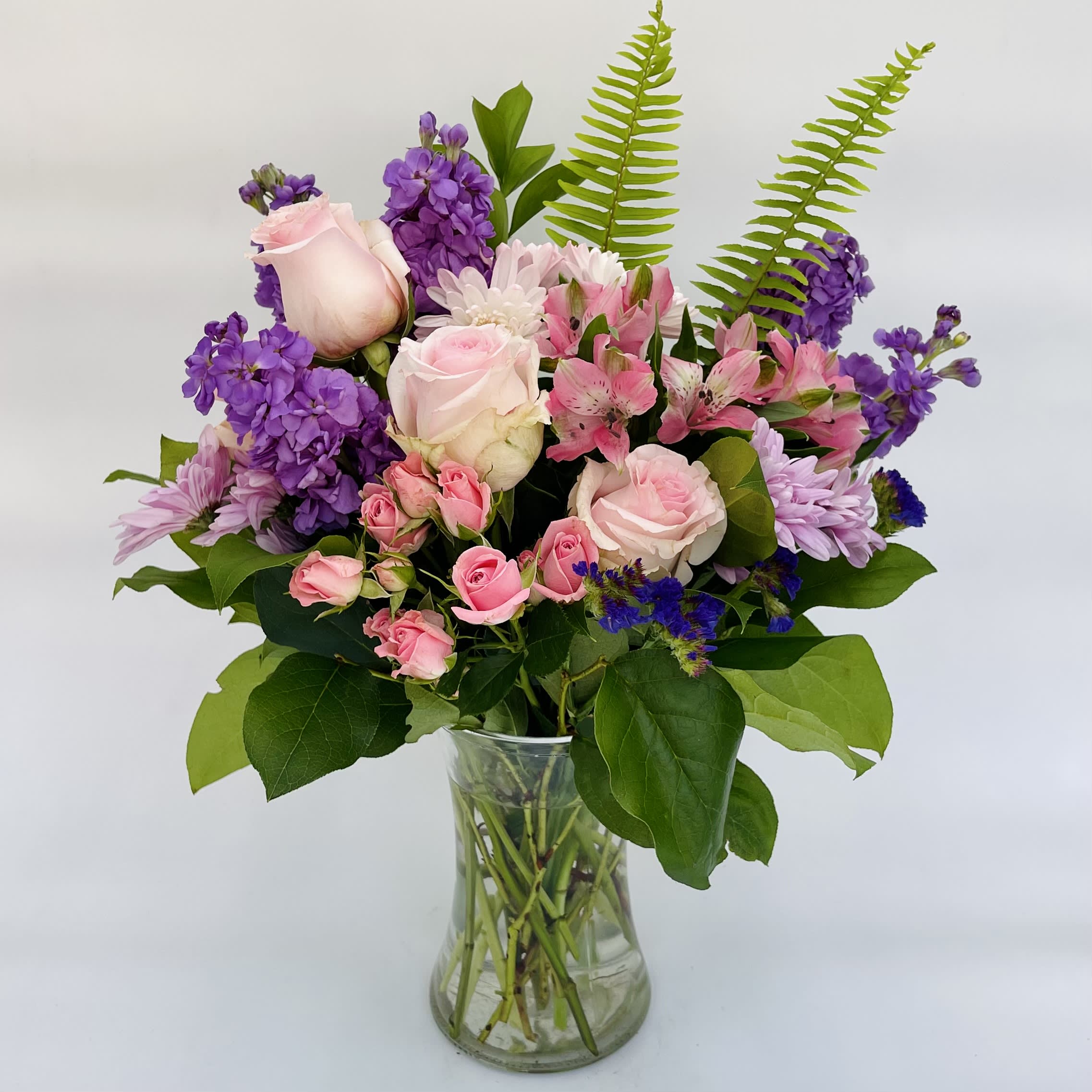Mother's Day Purple and Pink Clear Glass Vase - A bouquet of purple and pink blooms in a clear glass vase is a perfect gift for any occasion.  Arrangement includes pink roses, alstromeria, lavender daisy poms, purple stock and statice, accented with greenery.