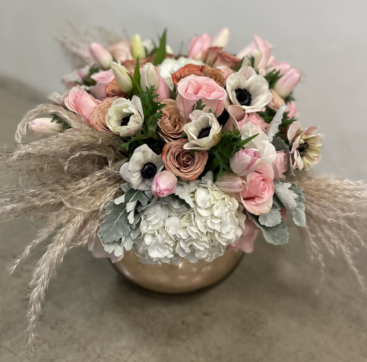 Emma - A muted pallete of roses, tulips, hydrangeas, anemones, dusty miller and pampas grass in a rose gold glass vase in 8&quot;D. Arrangement stands approximately 12&quot; high