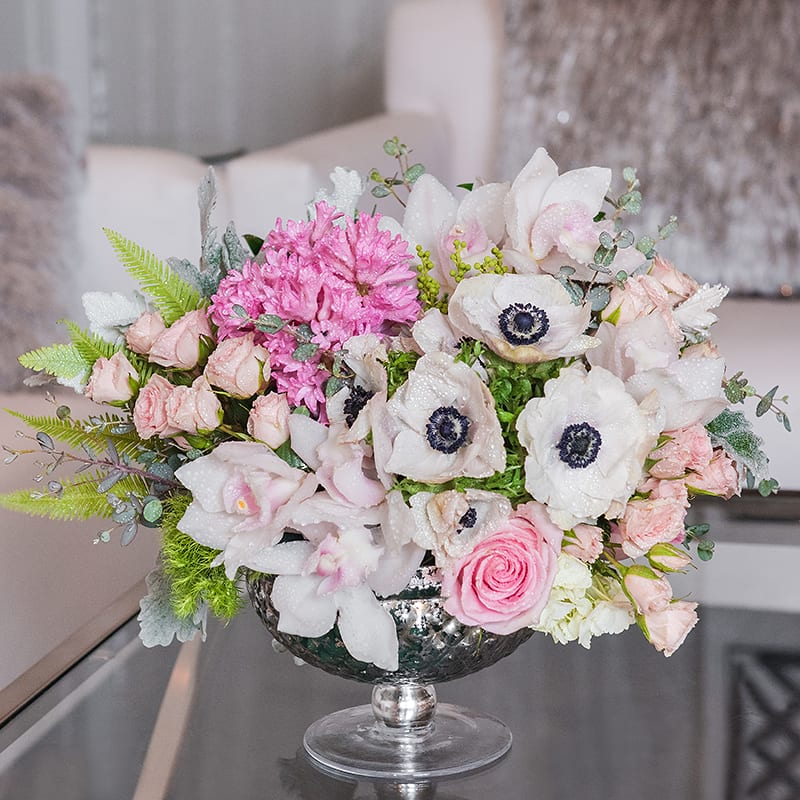 Blush Garden - This garden-style arrangement that's full of soft blush and pink flowers in an antique silver pedestal vase. A fragrant and darling gift overflowing with anemones, roses, spray roses, hyacinth, and cymbidium orchids.