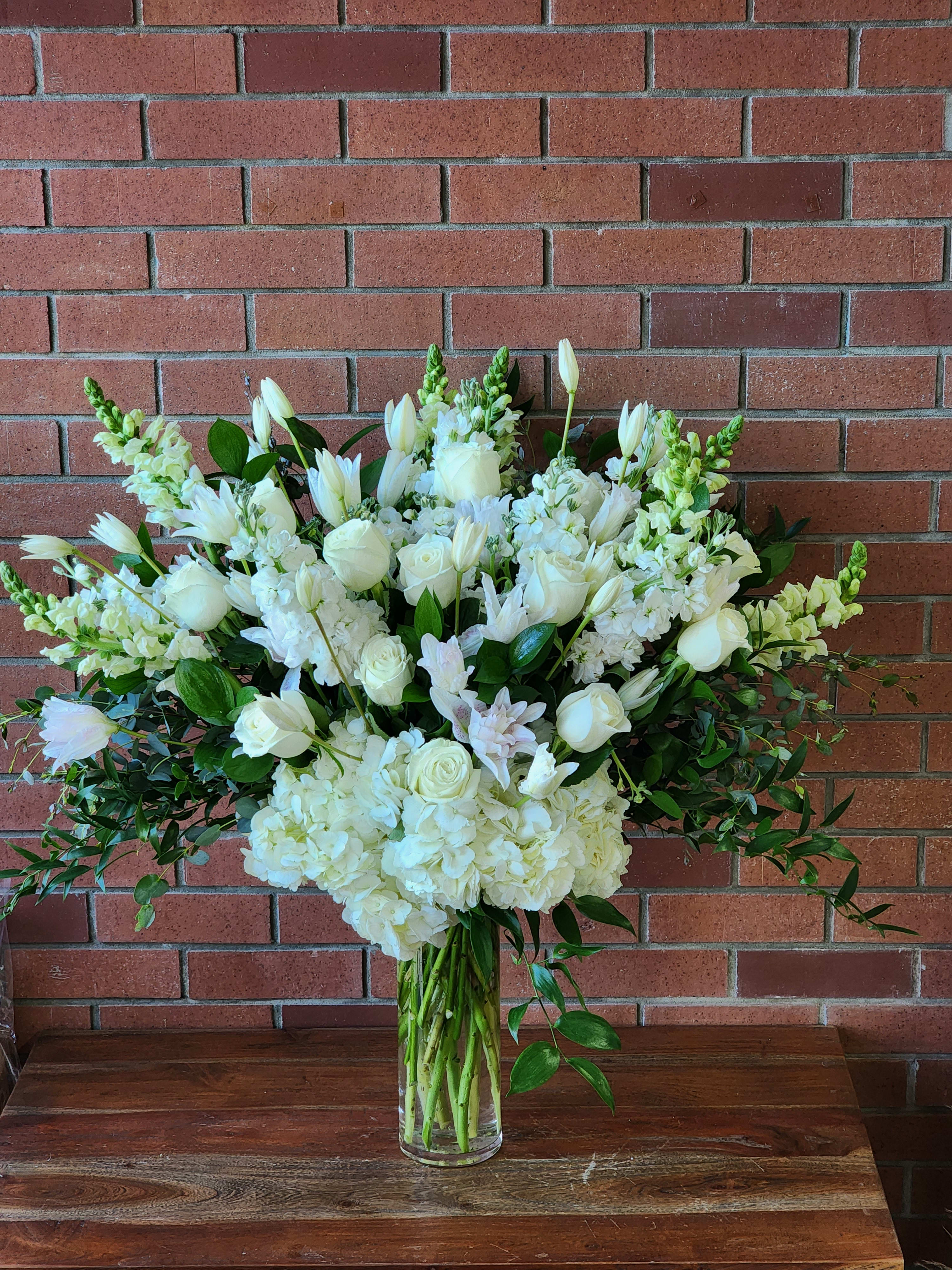 Large White Sympathy Vase Arrangement Large White Sympathy Vase Arrangement - Send your most sincere condolences and offer comfort and beauty in times of sorrow with our elegant all-white sympathy arrangement. White roses, lilies, stock, Eucalyptus, snap dragons Italian ruscus and hydrangeas in a clear tall cylinder vase.