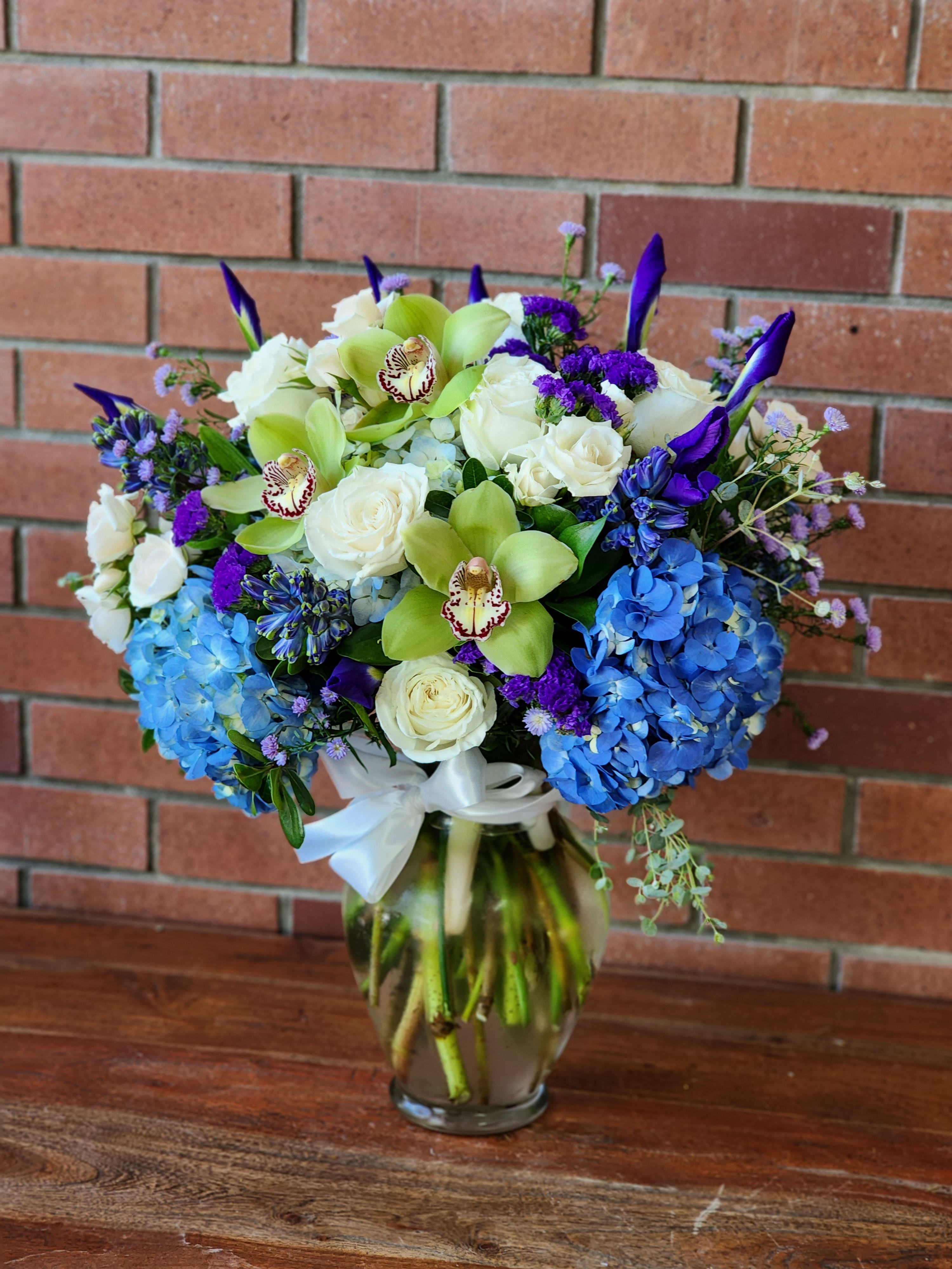 Moody Blues Bouquet Moody Blues Bouquet - This beautiful arrangement is sure to compliment anyone's special day or occasion. Blue hydrangeas, blue irises, white roses, spray roses, blue hyacinths and green Orchids. Arranged in a pretty rose vase with a silk white ribbon.