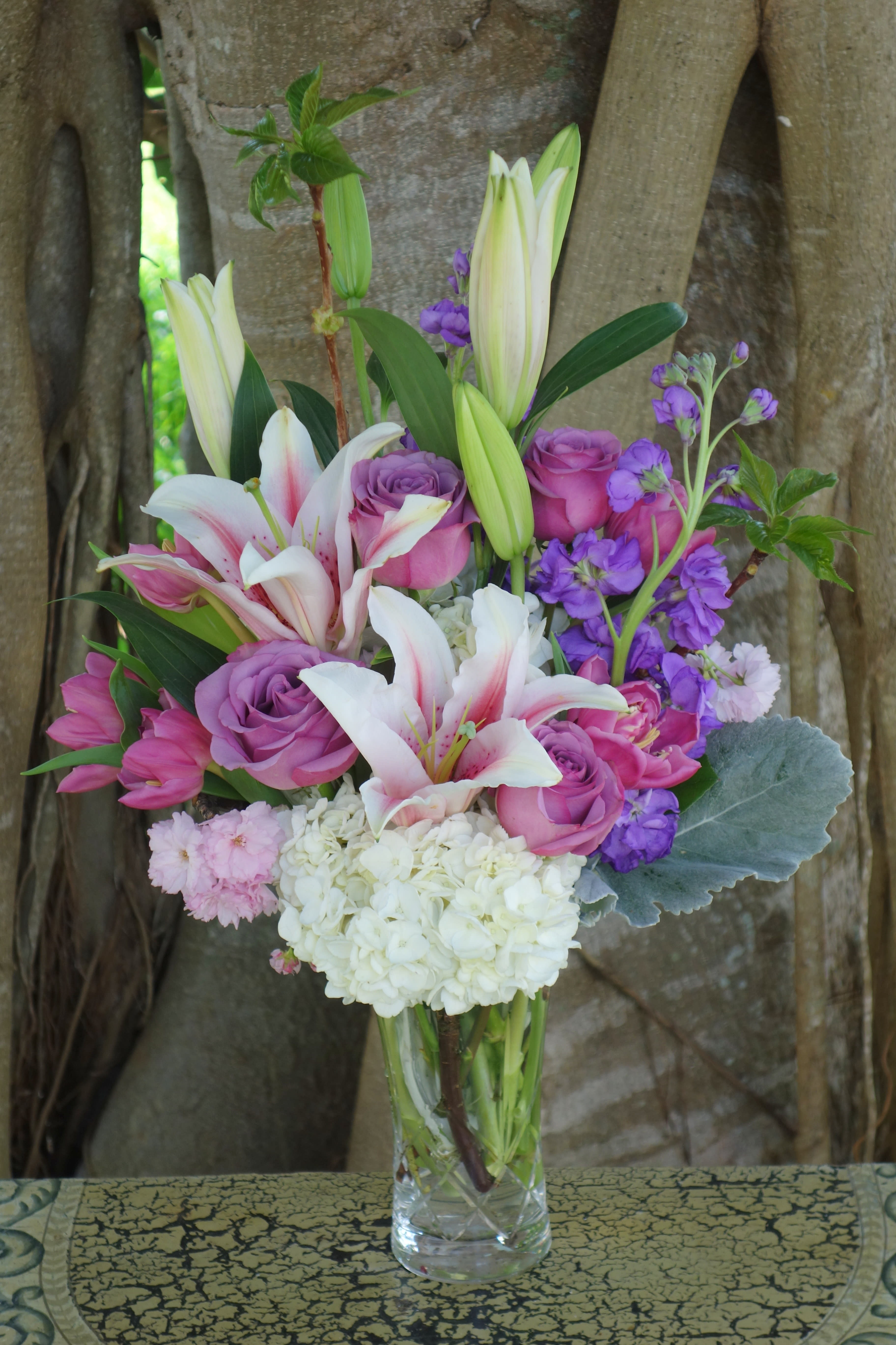 Elegant Wishes Elegant Wishes - Fragrant lilies, stock, cherry blossoms, purple roses, hydrangeas fillers and greens in a classic heavy glass checkered vase.