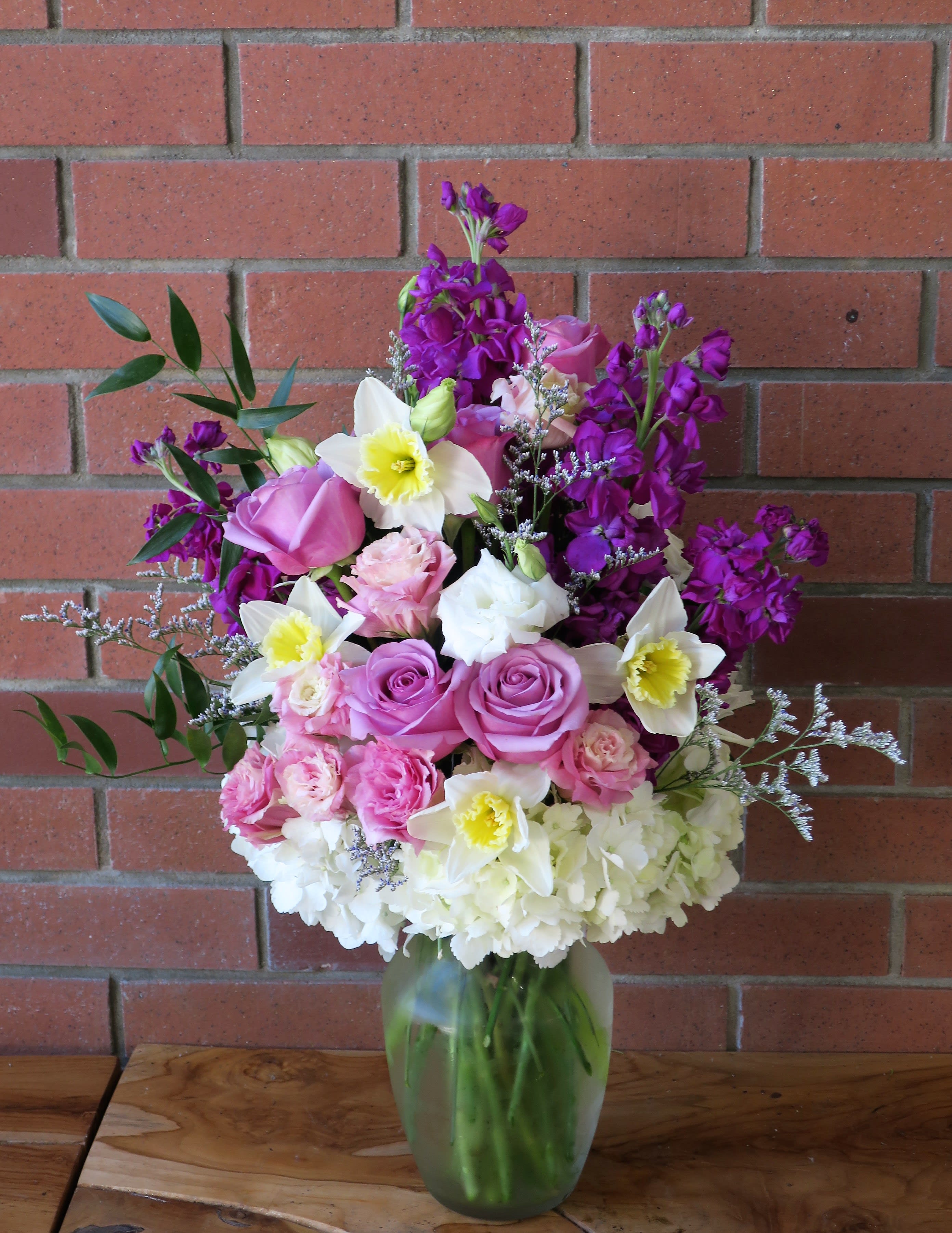 Signs Of Spring Signs Of Spring - Beautiful spring bouquet with fragrant stock, daffodils, purple roses, hydrangeas, fillers and greens in a clear vase.