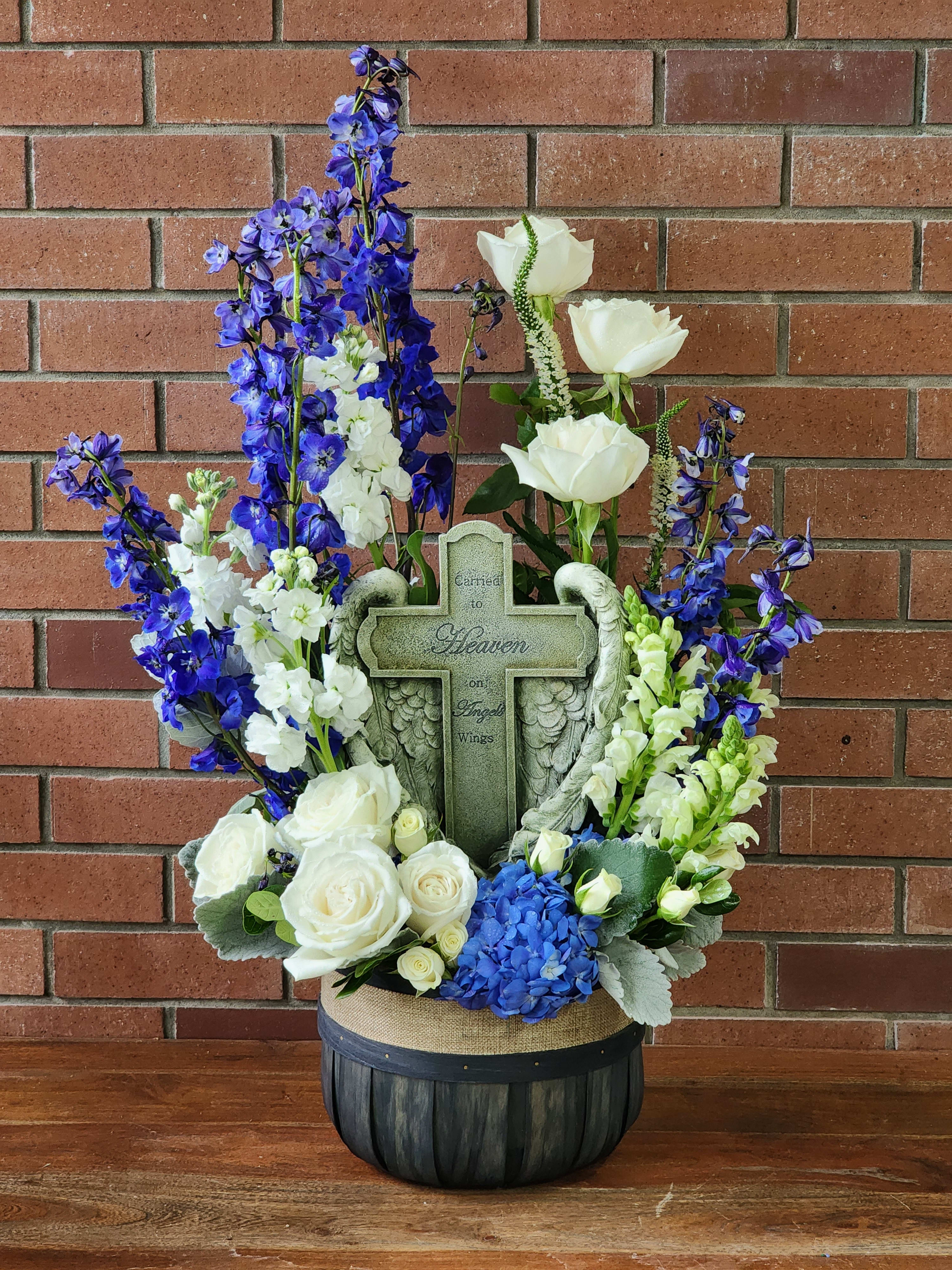 Carried To Heaven On Angel's Wings  - A beautiful keepsake cross hugged by angel wings sits in the middle of a high quality basket filled with blue delphinium, white roses, white stock, white spray roses, white snap dragons, greenery and dusty Miller. This basket is large. Approximate height is about 32&quot; by 18&quot; but sizes may vary based on the flowers put into the arrangement.