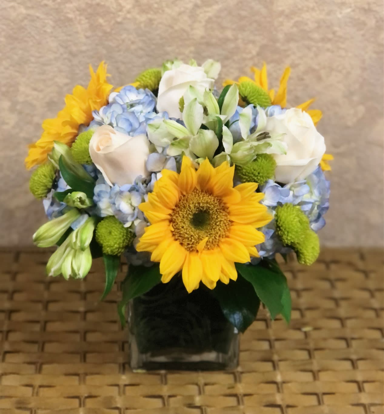 Sunshine by the Ocean - Our most selling bouquet is a summer gift everyone will love with ocean blues and yellow sunny sunflowers. Type of flowers: Blue Hydrangeas, Sunflowers, White Roses, White Alstroemerias and Green Button Mums in a clear vase with a deep blue wrap. Season Availability: All year round Dimensions: Square clear vase 5&quot; by 4&quot; Substitute Availability: Yes