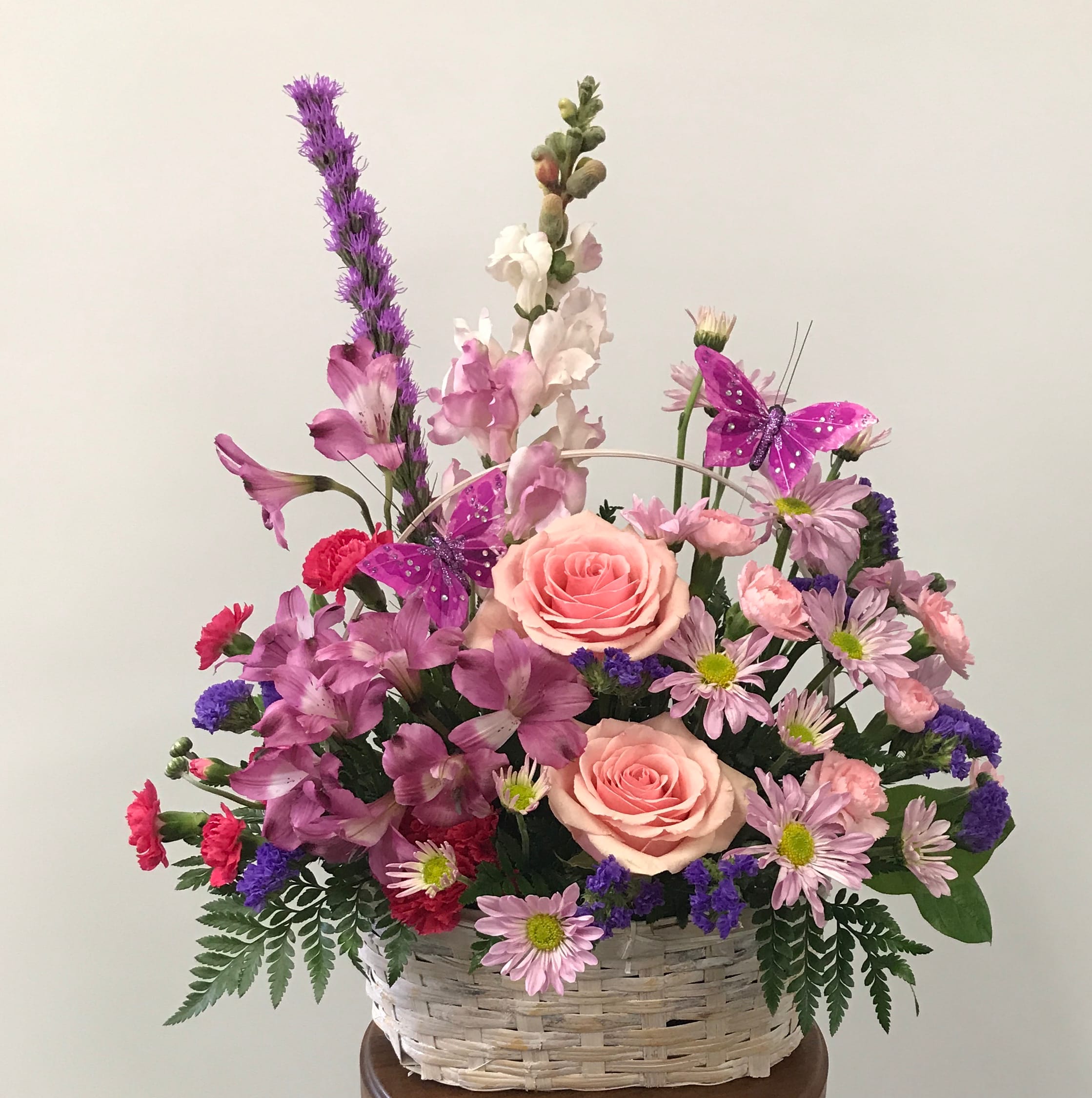 Country Garden Blooms - Talk about a bountiful bouquet! This basket is overflowing with beauty and blossoms. It's no wonder two pretty butterflies have made this arrangement their home. Flowers SUCH AS pink roses, alstroemeria, snapdragons and miniature carnations, purple daisies and statice are used. You've got this gift handled!   Orientation: One-Sided.  Basket may vary.   