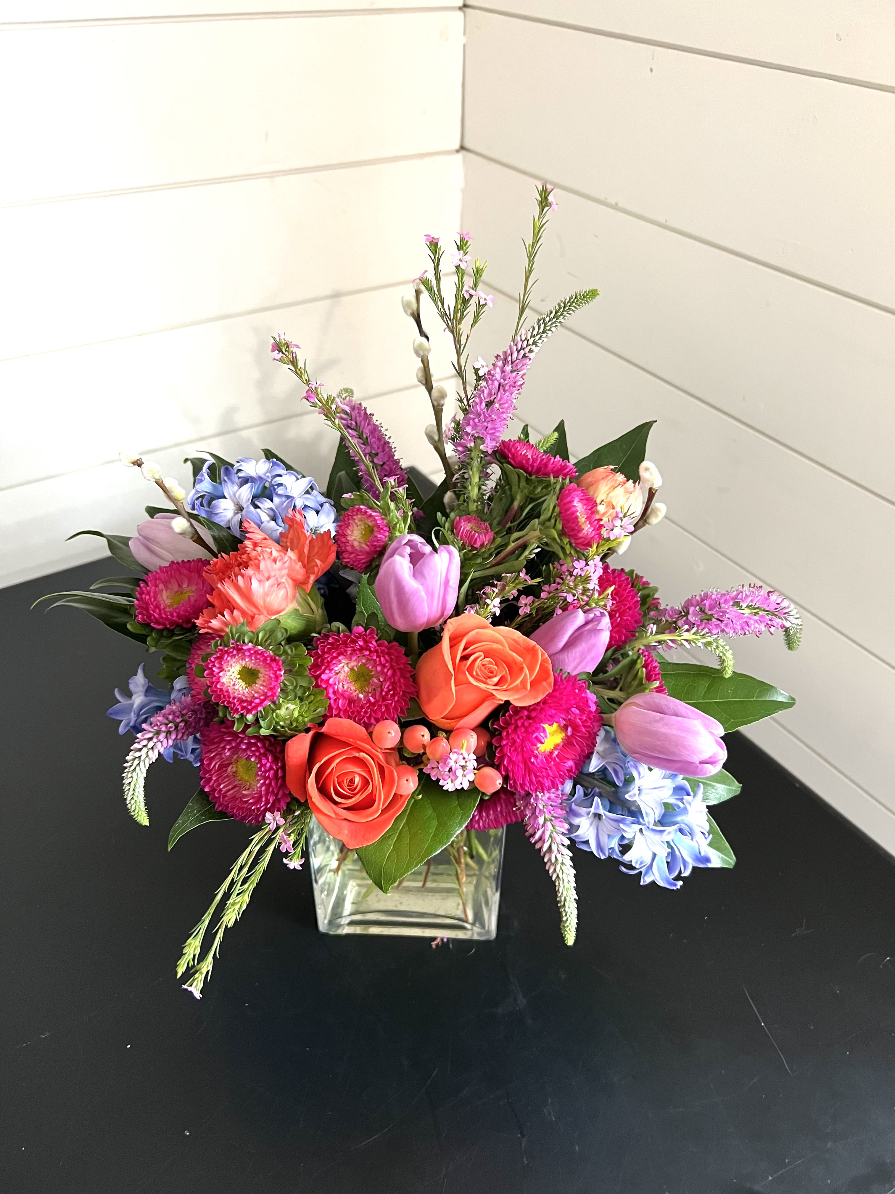 Pops of Color Arrangement - A lovely arrangement bursting with color and joy! A mixture of roses, tulips, and seasonal flowers on a bed of greenery in a clear vase.