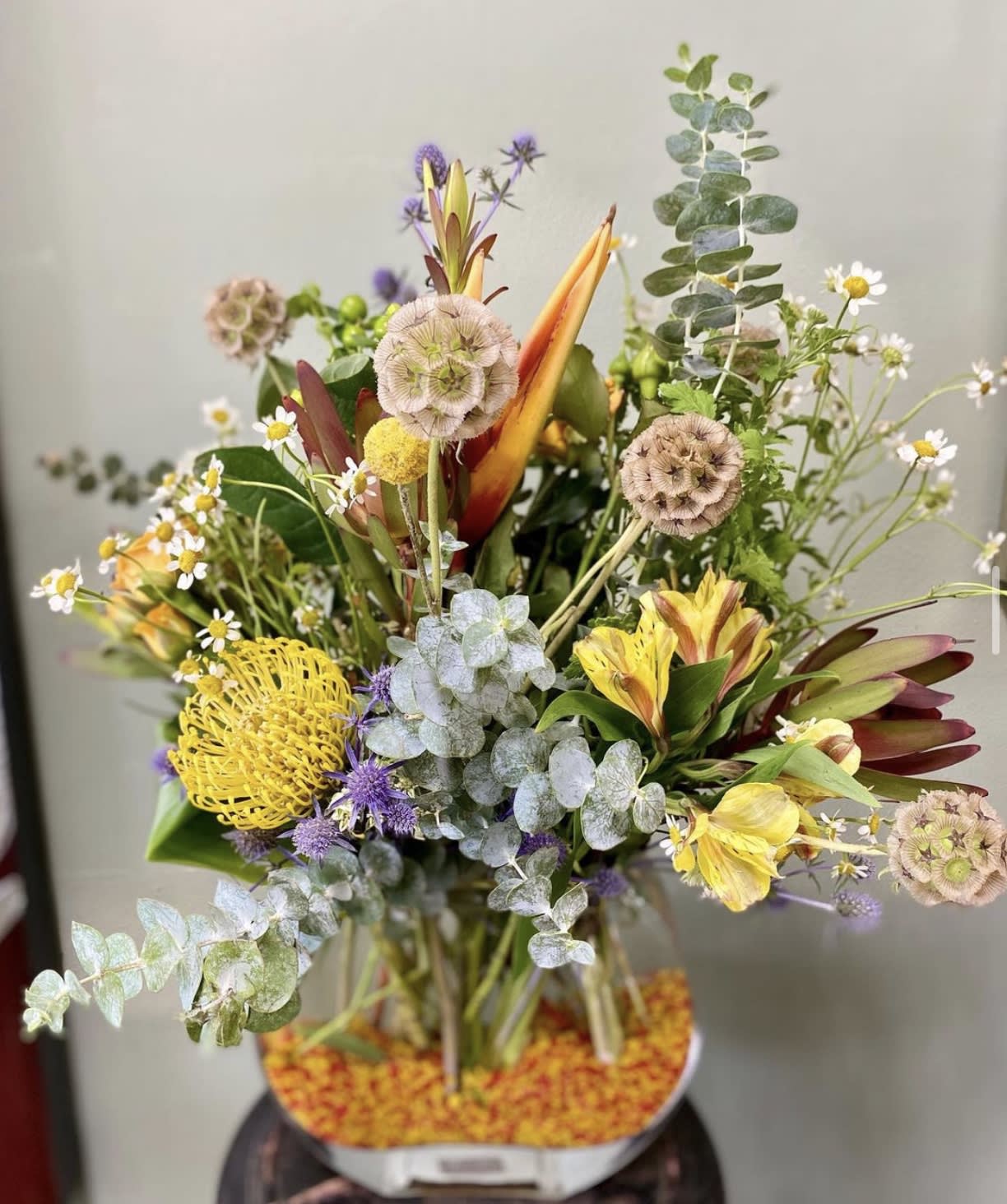 The Wild Yellow The Wild Yellow - Yellow Pincushion Protea, bird of paradise, coffee beans, blue teasel, yellow alstroemeria, yellow roses, eucalyptus, green filler etc, all hand arranged in glass vase.
