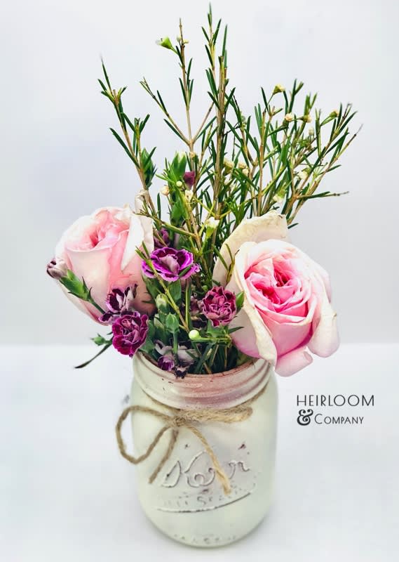 Rosey Mason Jar - This adorably rustic design is a favorite for anyone with a fun loving spirit and a love for anything farmhouse chic. Roses, and was wildflowers mixed in with baby carnations or mini spray roses presented in a blush handprinted mason jar.