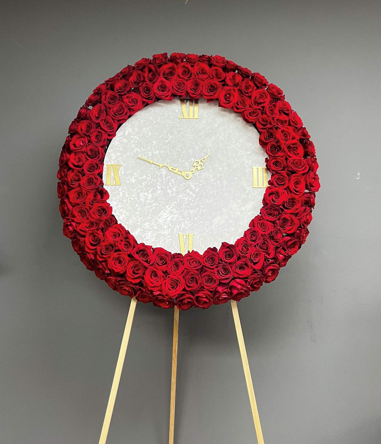Memorial frame with a stopped clock 2 - The memorial round frame is a poignant and symbolic tribute, designed to honor and remember a loved one who has passed away. It features a stopped clock as the centerpiece, surrounded by a bed of red roses and displayed on an easel. Additionally, a white or black ribbon with professionally printed letters is incorporated into the design. The round frame serves as a symbolic representation of the eternal nature of the departed soul. The stopped clock signifies that time has ceased for the individual, emphasizing the finality of their passing. It serves as a reminder to cherish the moments spent with the person and reflect on the preciousness of time. The red roses encircling the clock hold deep significance in the language of flowers. They symbolize love, respect, and courage. The vibrant red color signifies the intensity of emotions and the enduring love that continues even after death. The presence of the roses creates a visually striking contrast against the clock, adding a touch of warmth and beauty to the memorial arrangement. The addition of a white or black ribbon with professionally printed letters adds a personalized touch to the memorial arrangement. The ribbon can bear a name, a meaningful quote, or any other short text. For longer texts you can choose to attach printed sympathy card.  The choice of white or black ribbon depends on the desired aesthetic or symbolism. White often represents purity, peace, and spirituality, while black can convey solemnity, reverence, and mourning.