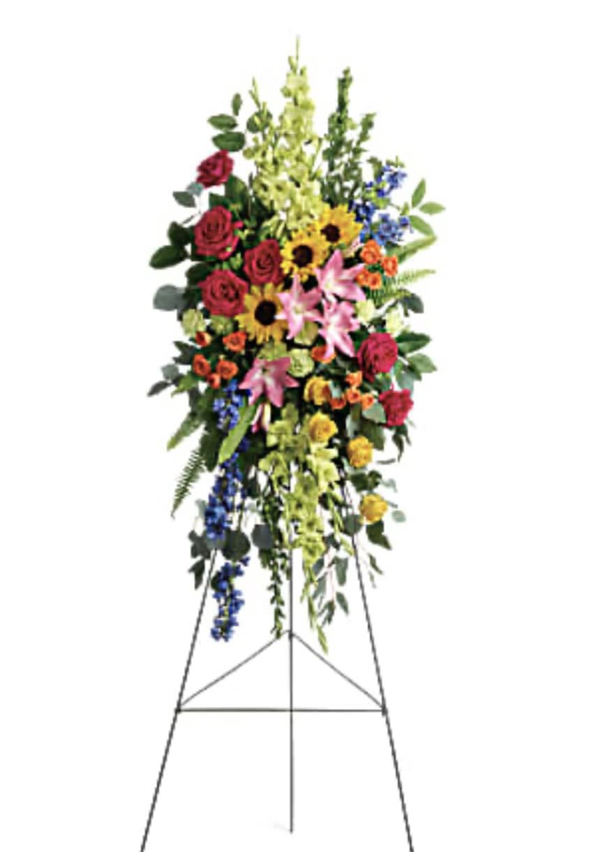 Love Lives On Spray - A bold, bright tribute, this rainbow of blooms fills the room with a spirit of hope and joy. What a glorious celebration of a love that will live on forever. This spray features hot pink roses, yellow roses, orange spray roses, pink asiatic lilies, green gladioli, green carnations, medium yellow sunflowers, blue delphinium, bupleurum, myrtle, sword fern, silver dollar eucalyptus, and lemon leaf.