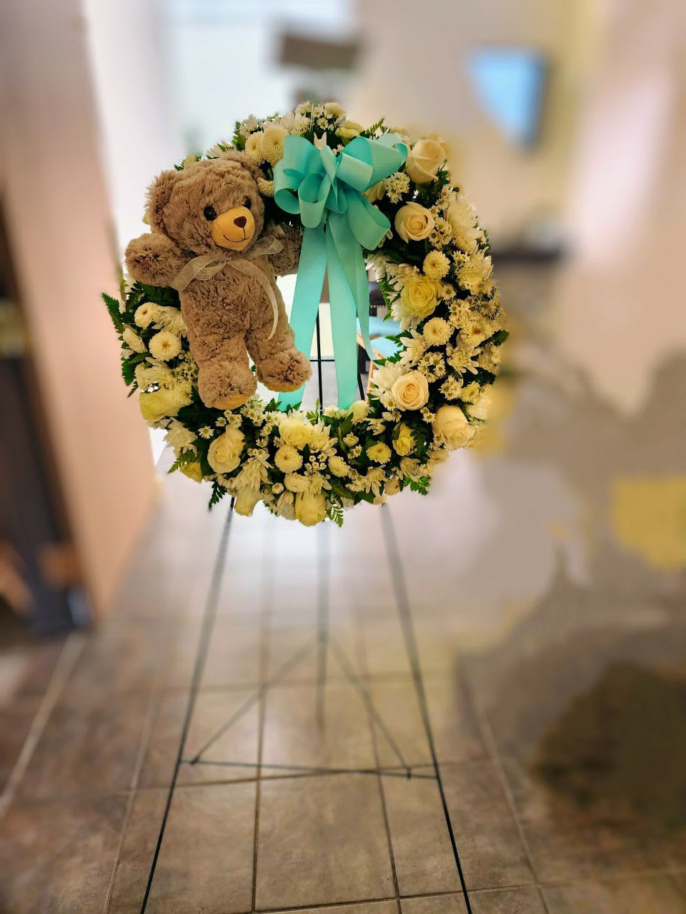 My Little Angel - A  very special detail  for a little angel .funeral wreath  has  12 white roses , 5 white cushion ,3 white  button ,3 white monte casino and a small brown  teddy  bear . With a final touch of a light blue bow in the middle of the arrangement  Comes with a wooden easel. the  wreath has a approximate measure of  18 inches 